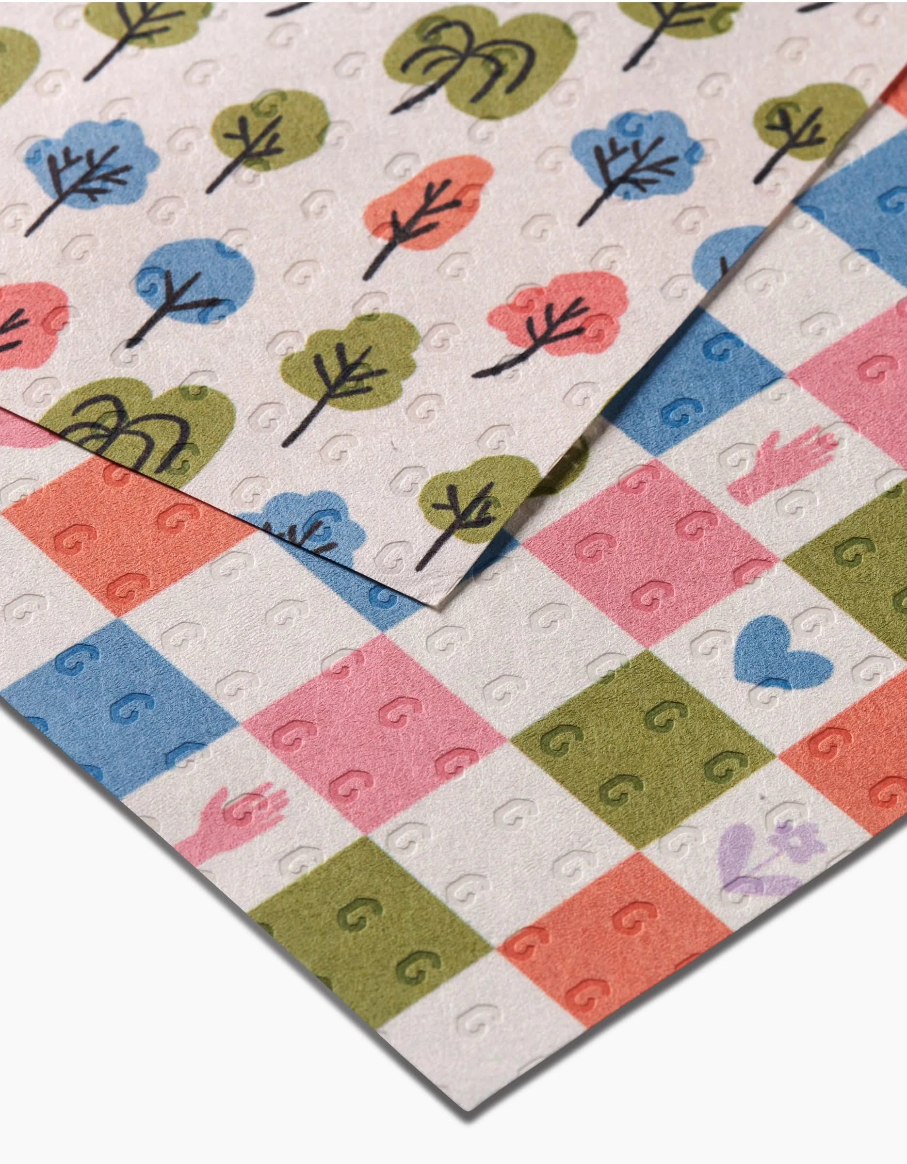 Two patterned papers overlap: Kayla Ann’s Happy Checkers, with animal and heart motifs in a checkered design, and another eco-friendly sheet made from post-consumer recycled materials, printed with colorful trees and black trunks.