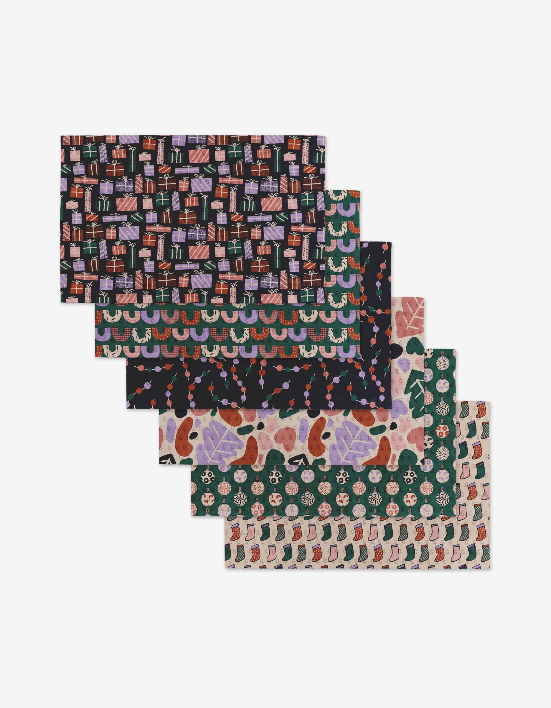 Six rectangular sheets of Happy Holly by Claire Iglesias are arranged in a staggered stack, each with festive patterns like gifts, garlands, gingerbread, snowflakes, and stockings—made from eco-friendly post-consumer recycled materials.