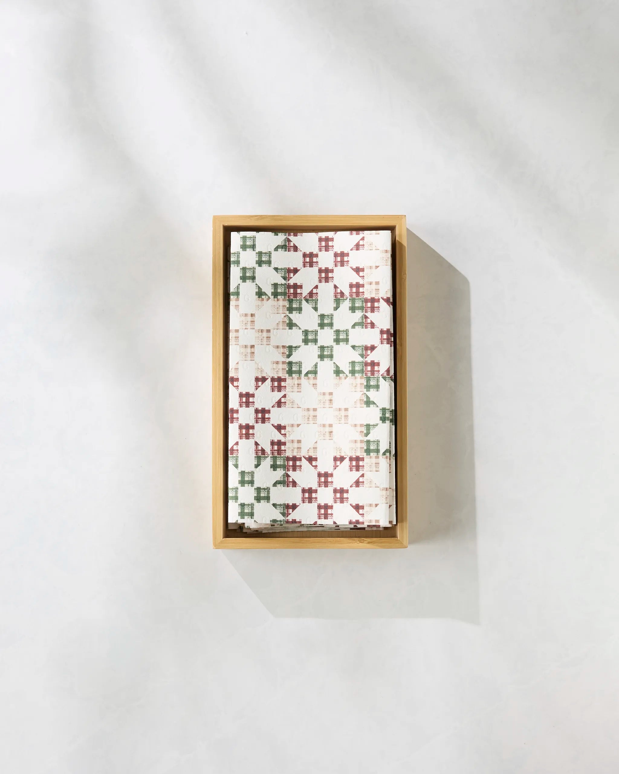A rectangular wooden tray holds a stack of Geometry’s Holiday Quilt eco-friendly paper towels, featuring a red and green geometric pattern, set on a light, textured surface with soft shadows.