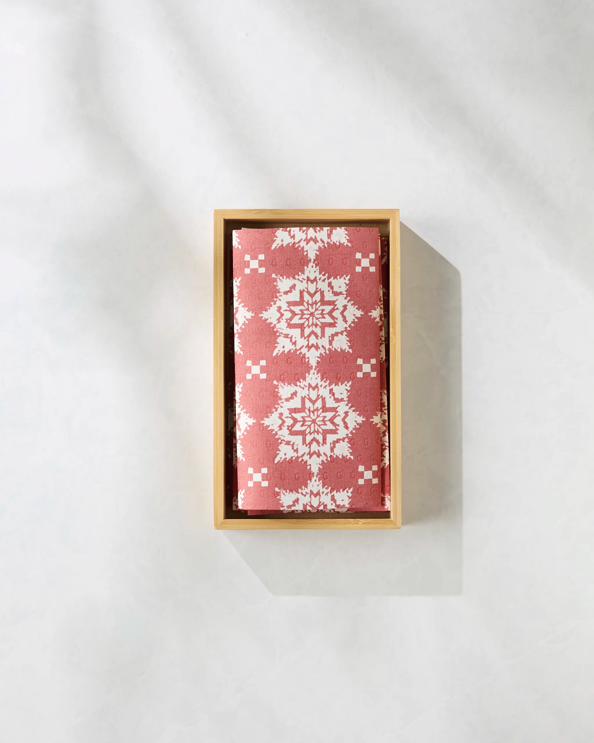 A rectangular wooden box holds a folded Geometry Iceflower Red cloth, featuring a white geometric and floral design, resembling reusable paper towels, displayed on a light textured surface with soft shadows.
