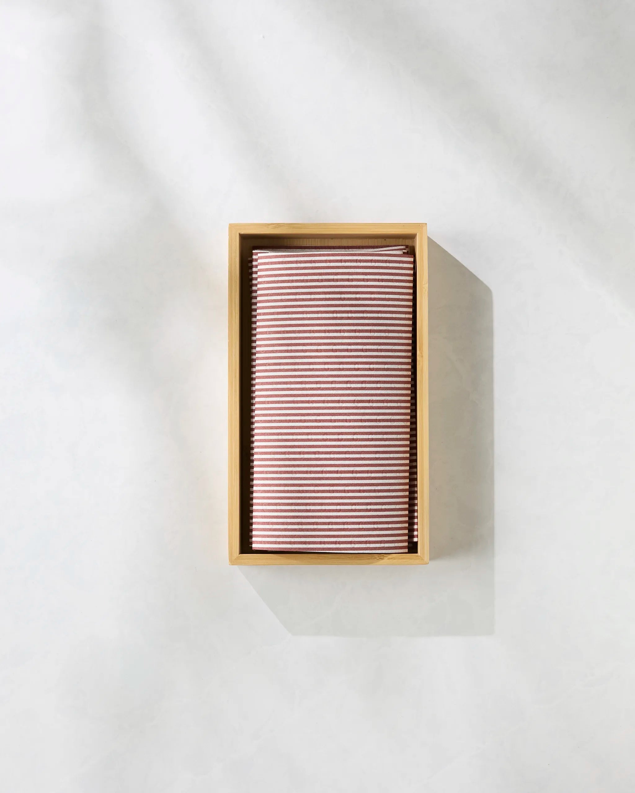 The Geometry Kringle kitchen towel, featuring burgundy and white stripes and made from eco-friendly materials, is neatly folded inside a rectangular wooden box against a softly textured light background with subtle shadows.