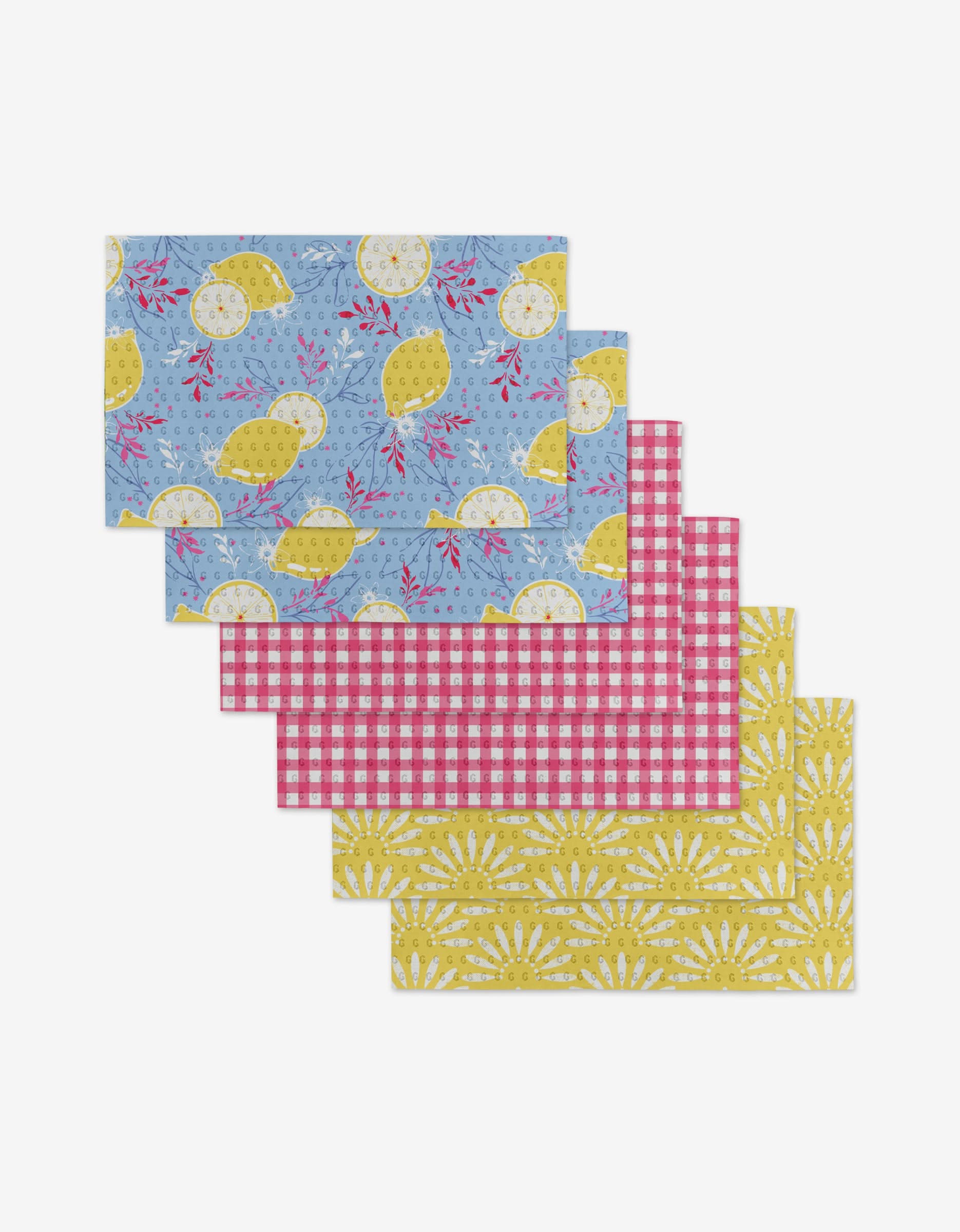 Ashley Celena’s Lemon Pop set offers six rectangular placemats in blue with lemons, pink and white gingham, and yellow floral sunbursts—an eco-friendly alternative to disposables, stylishly arranged in a fanned-out stack.