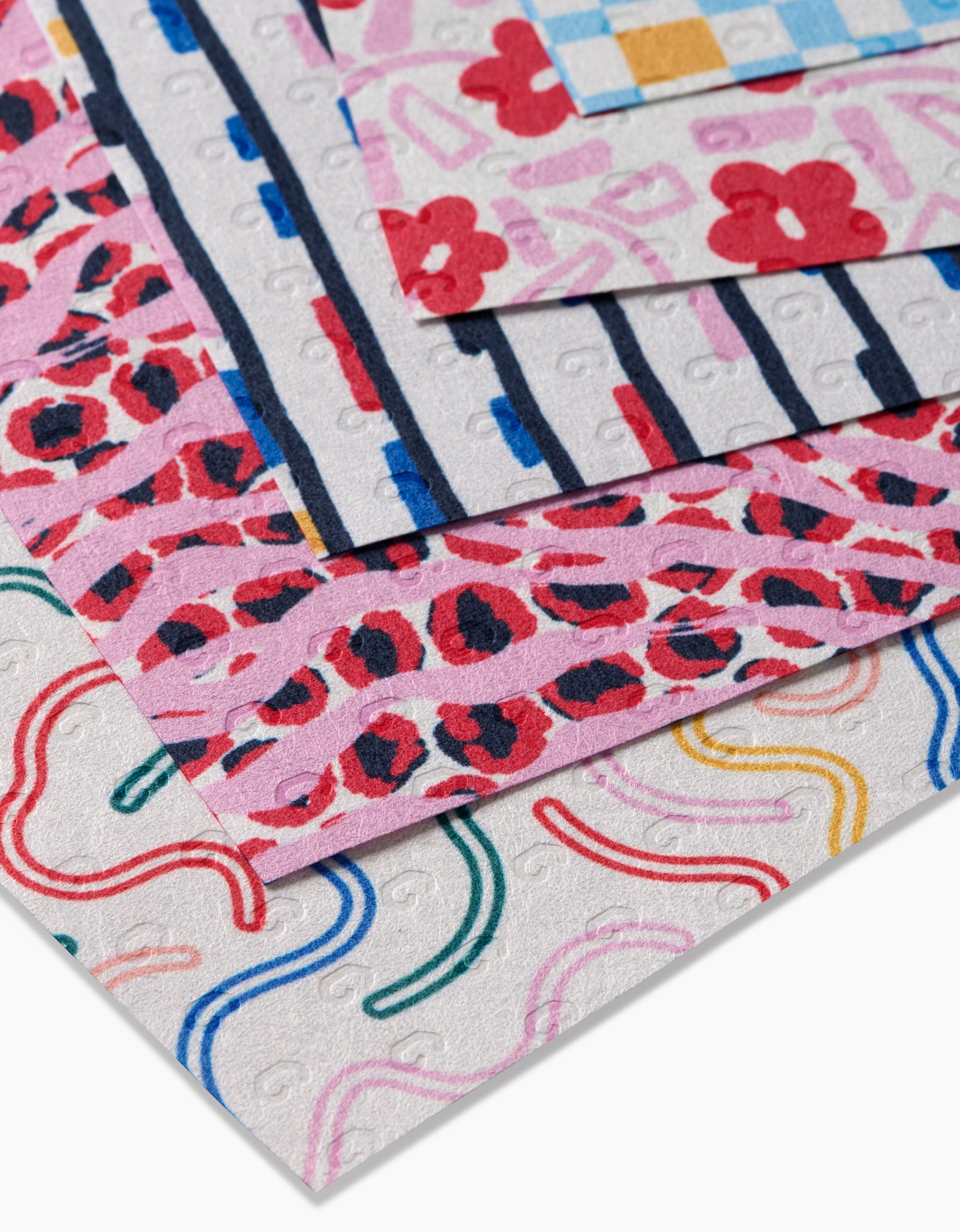 A close-up of stacked Magic Wand squares by Happy Hemera, made from recycled materials and featuring colorful floral, animal, striped, plaid, and curved designs on a white background.