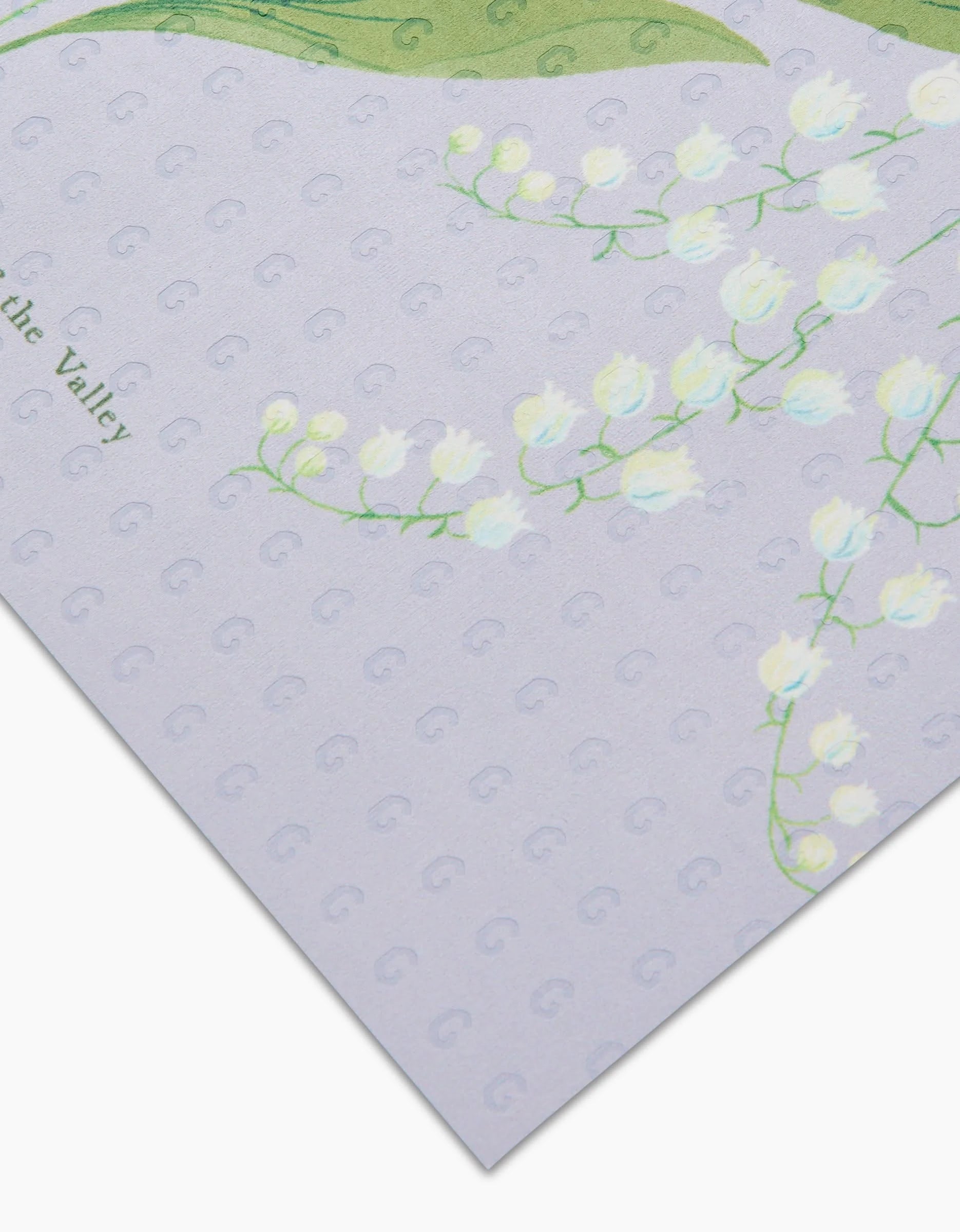 A close-up of Linda Fox’s May Lilly Of The Valley: an eco-friendly, reusable pastel sheet adorned with white lily of the valley flowers and green leaves over a light lavender dotted background—not paper towels.