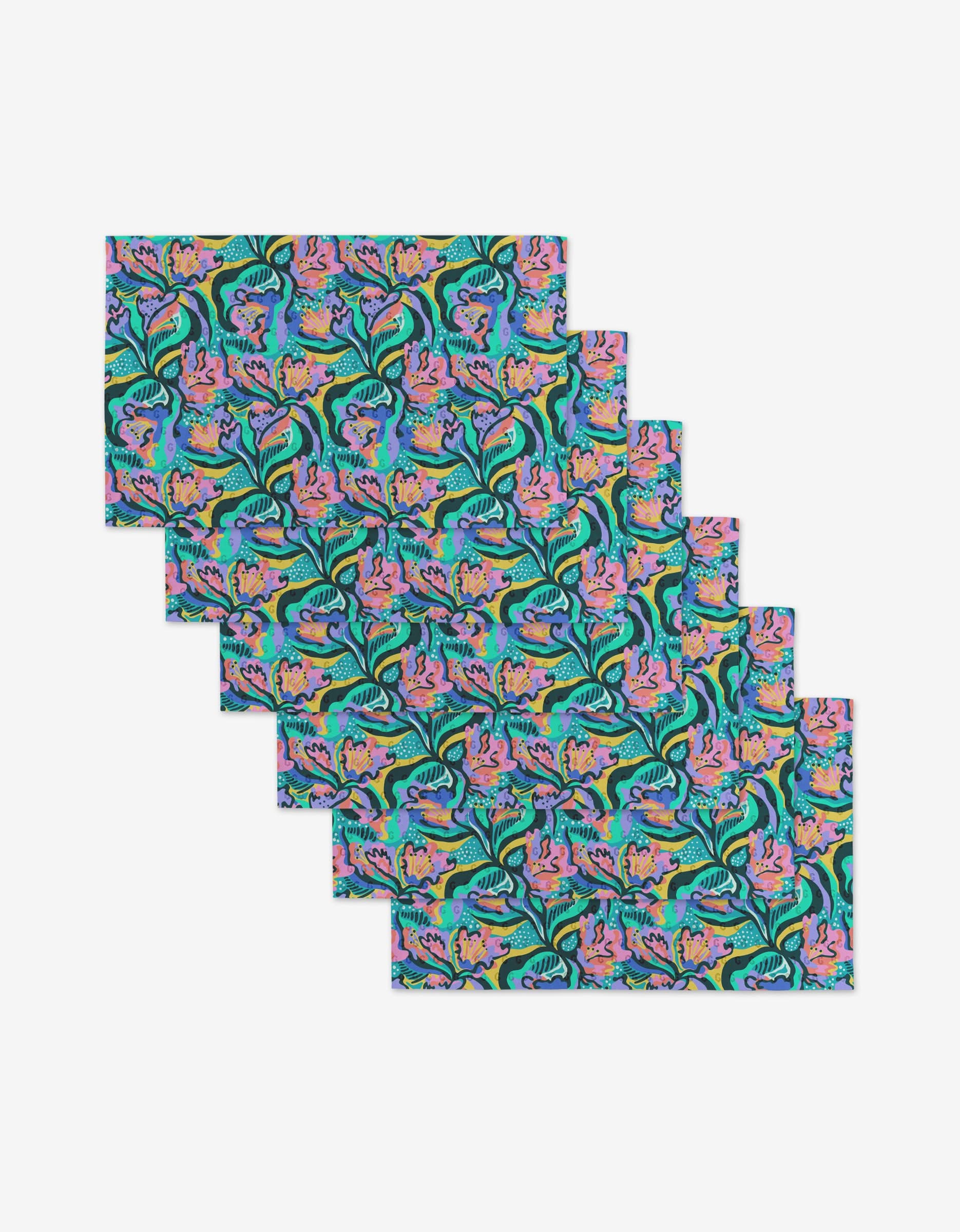 Six Moody Flowers rectangular placemats by Masha Volnova are stacked, each with a vibrant floral and leaf pattern in aqua, pink, purple, yellow, and green on a dark background—crafted from eco-friendly materials for sustainable style.