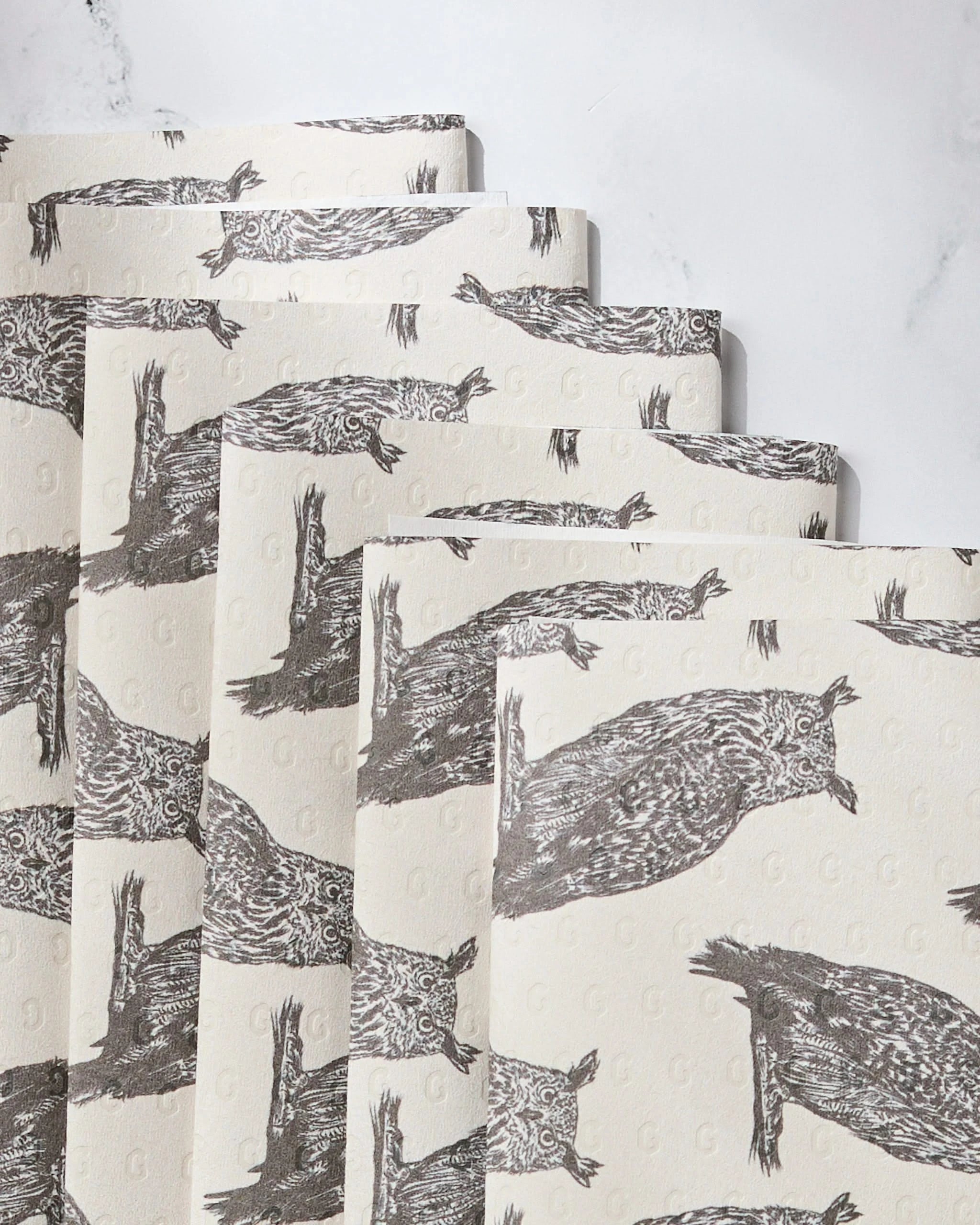 Five sheets of Geometrys Nocturne reusable paper towels, each featuring detailed brown owl sketches on cream, are stacked horizontally on a white marble surface.