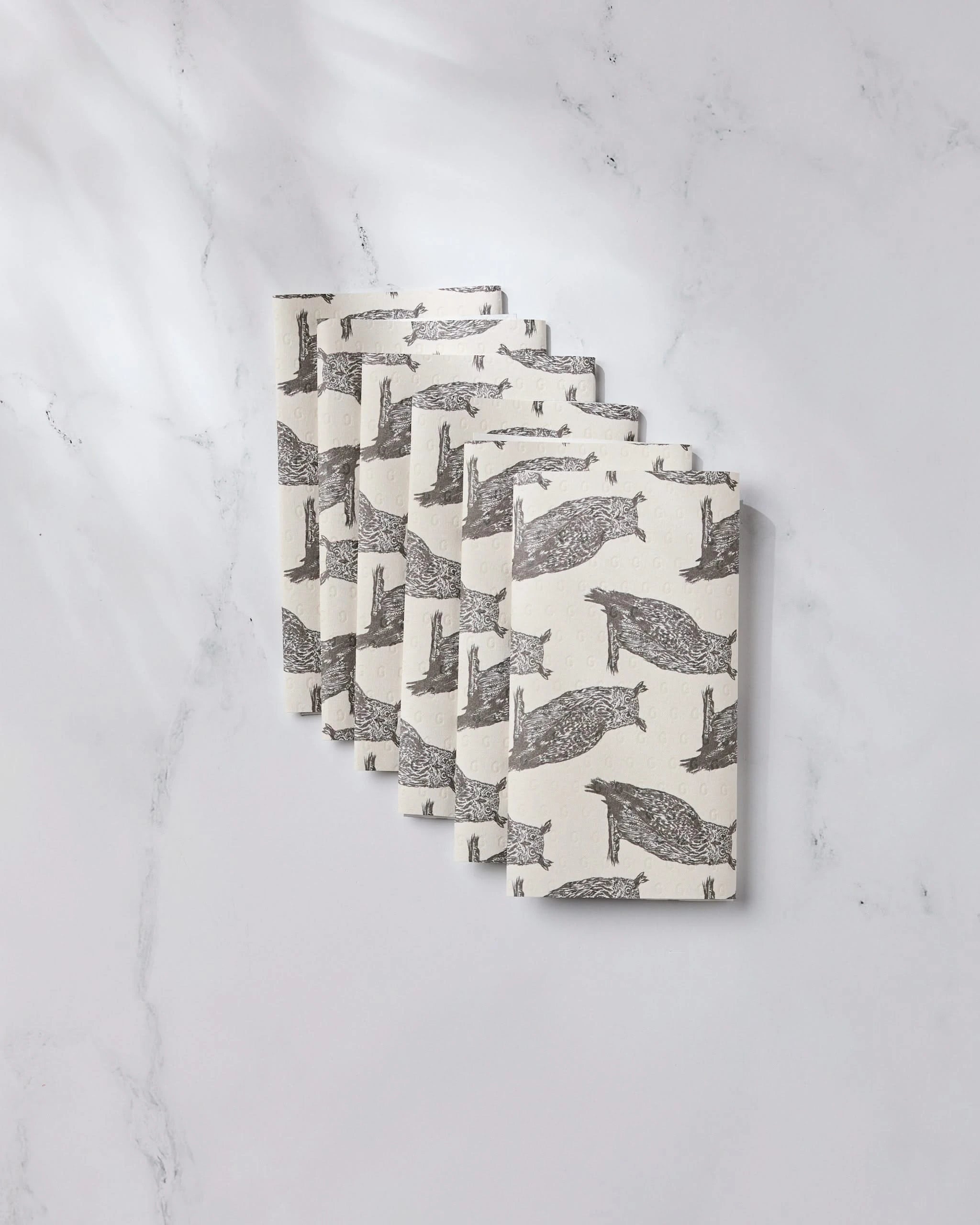 Seven Nocturne rectangular bars by Geometry, featuring a black fish pattern and resembling eco-friendly kitchen towels, are diagonally arranged and partially overlapping on a white marble surface.