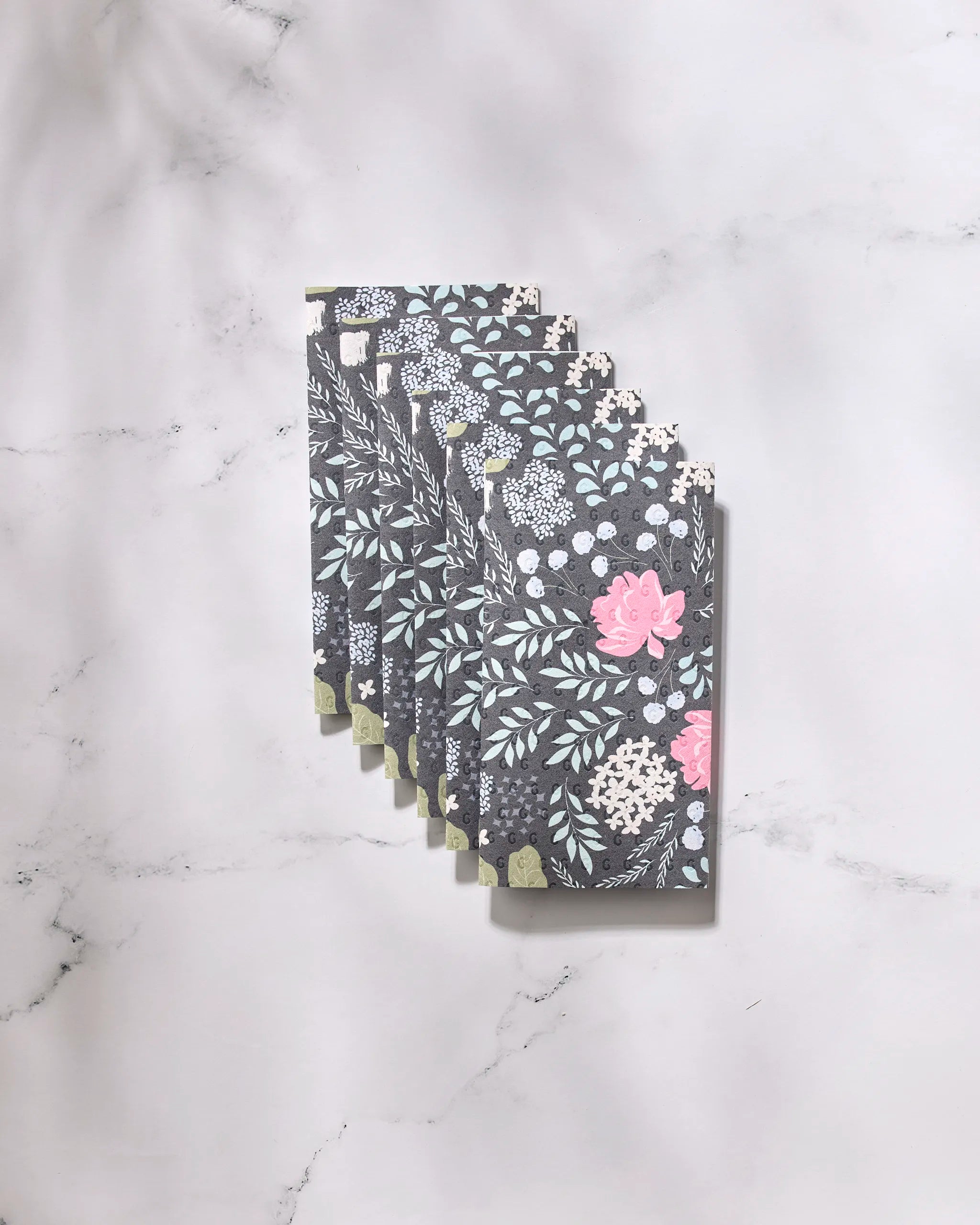 A stack of GEOMETRY Not Forgotten notebooks with dark, floral-patterned covers and pastel accents rests diagonally on a marble surface, accompanied by reusable paper towels for an eco-friendly touch.