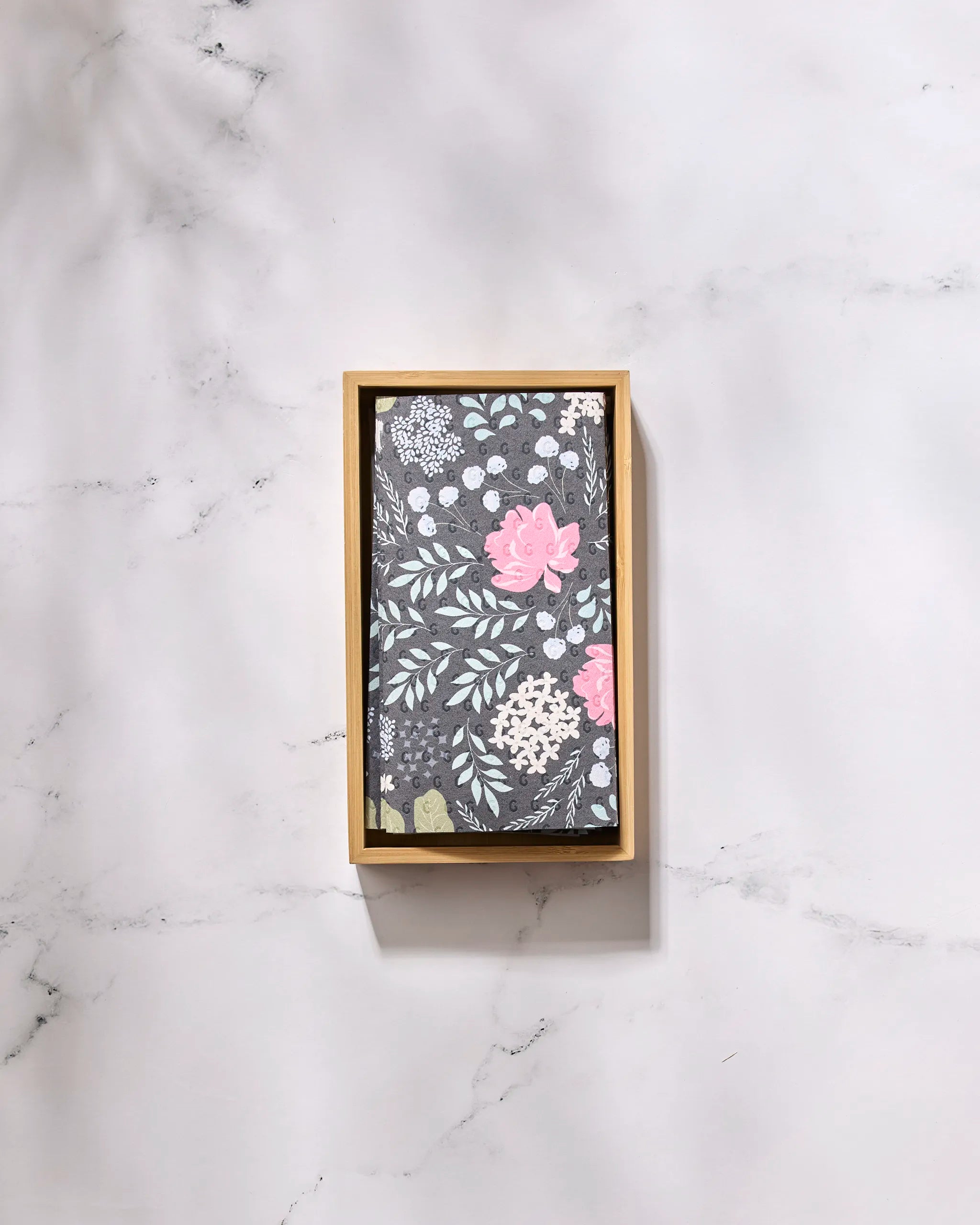 The Not Forgotten notebook by GEOMETRY, featuring pink, white, and green floral patterns on a dark cover, is placed in a rectangular wooden tray on white marble next to eco-friendly kitchen towels.