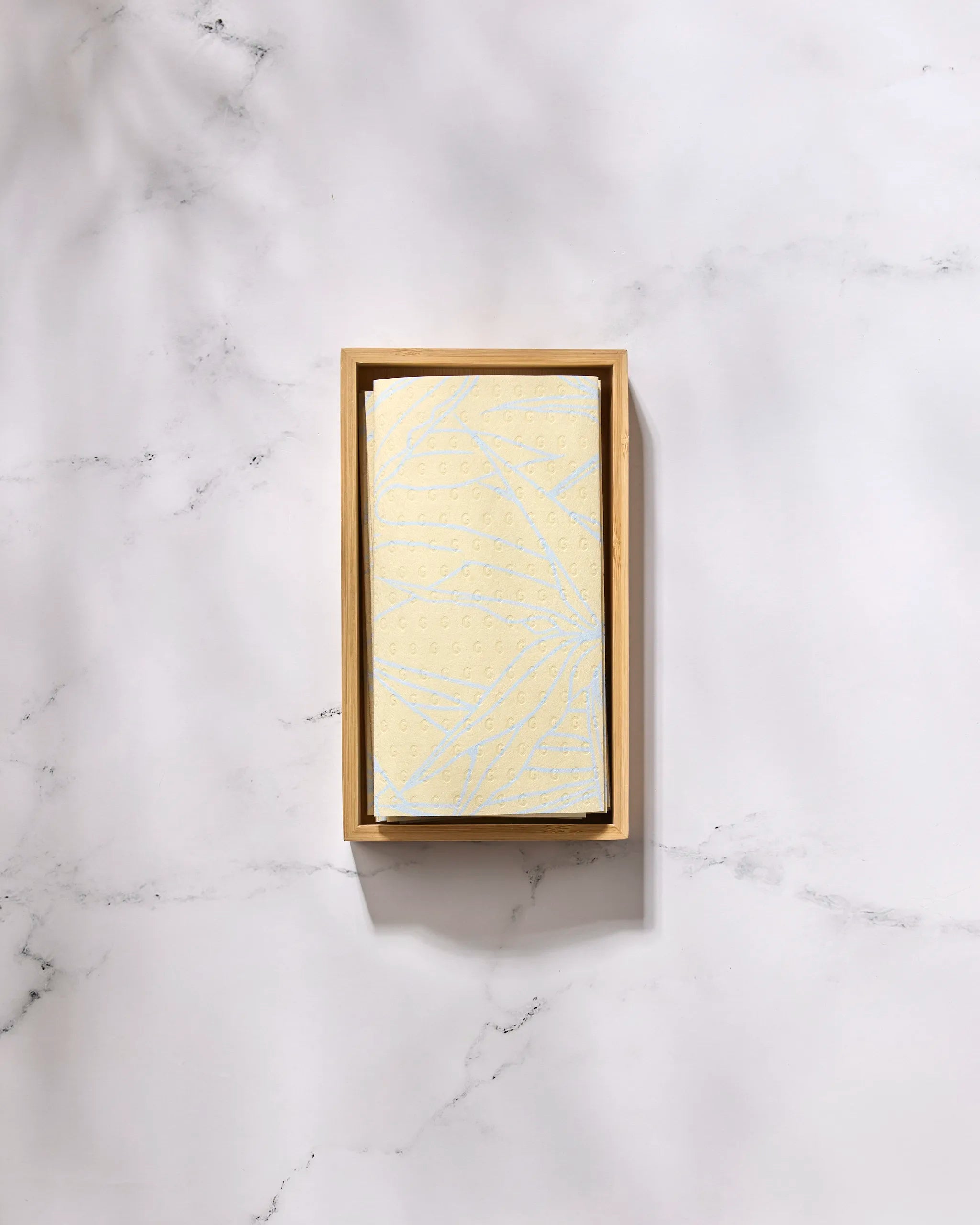 The GEOMETRY Paradise Blooms eco-friendly, pale yellow paper towel with a subtle pattern rests neatly folded in a rectangular wooden tray on a white marble surface.
