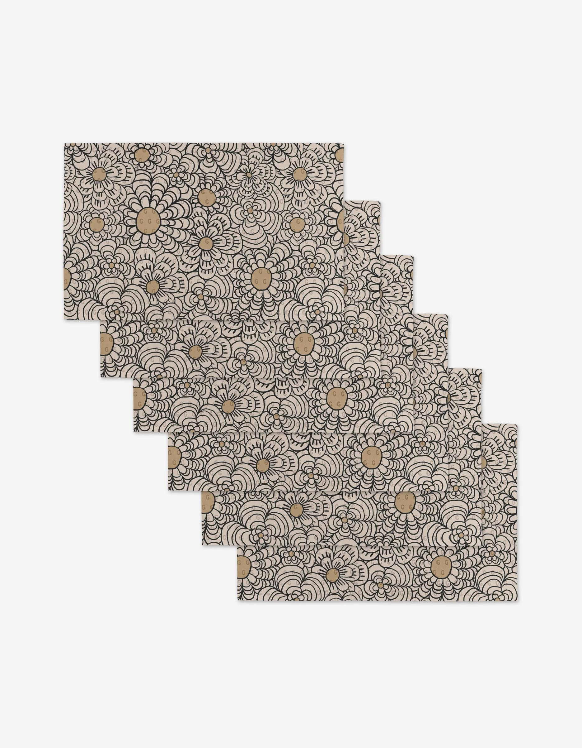 The Paulette placemat set by Amy MacCready includes six rectangular mats with beige backgrounds, black floral patterns, gold flower accents, and is made from eco-friendly post-consumer recycled materials.