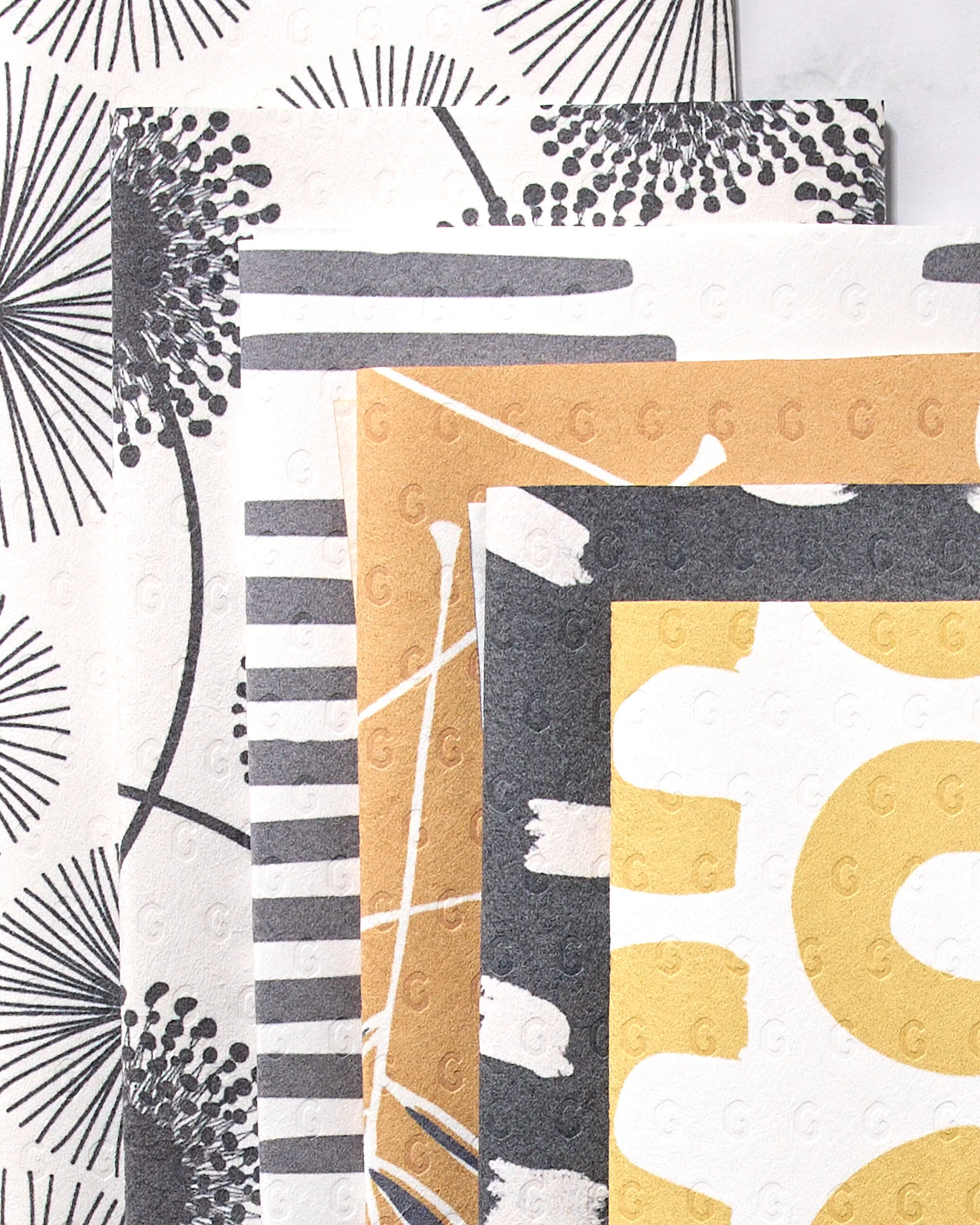 Four GEOMETRY Pop Of Color reusable towels, featuring bold modern patterns in black, white, gold, and gray with abstract florals, stripes, and brush strokes designs, are shown overlapping. Each towel has a subtle embossed texture.