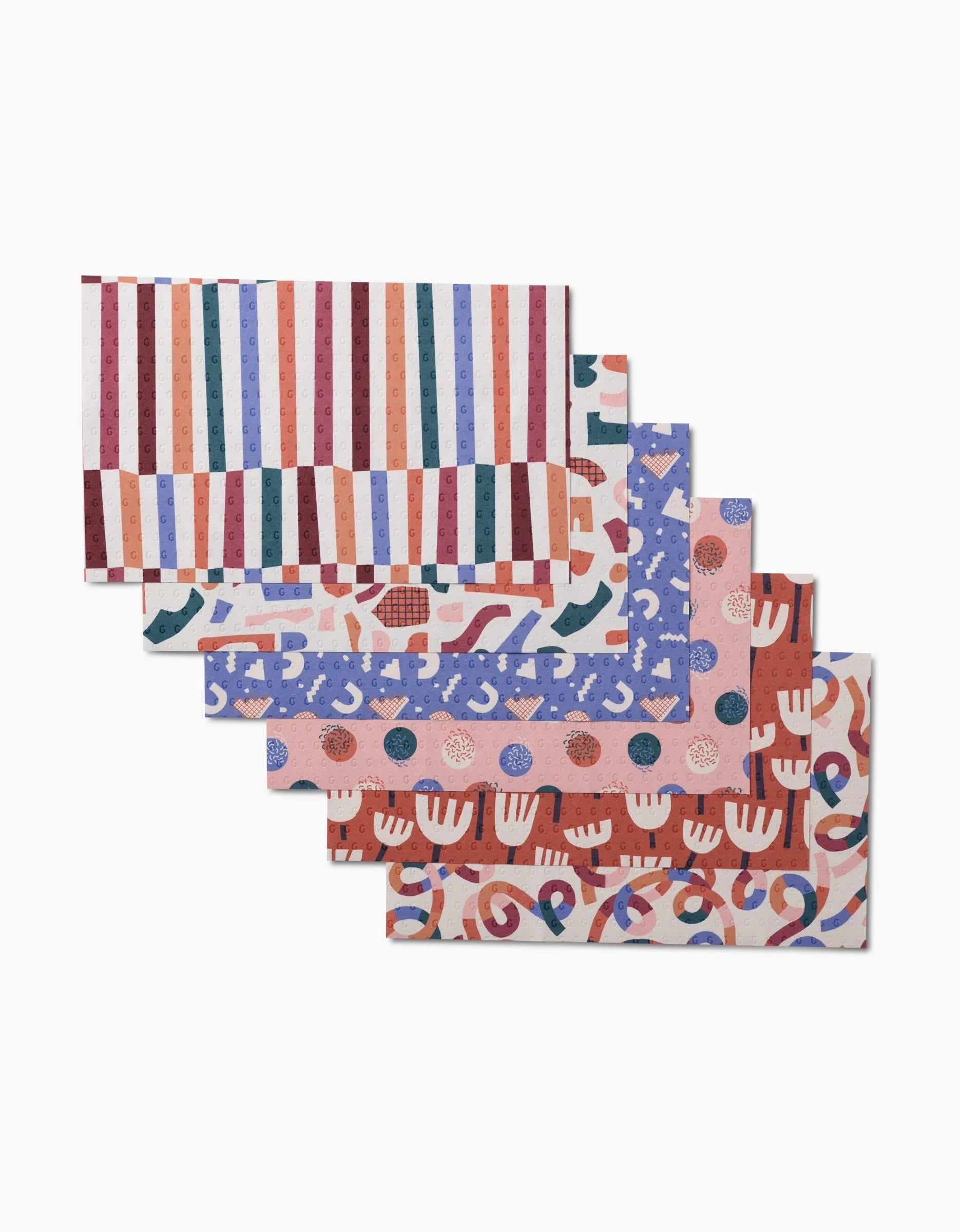 Five Rainbow Cutouts placemats by Claire Iglesias—rectangular and made from post-consumer recycled materials—feature colorful patterns including stripes, shapes, dots, hands, and squiggles fanned out on a white background.