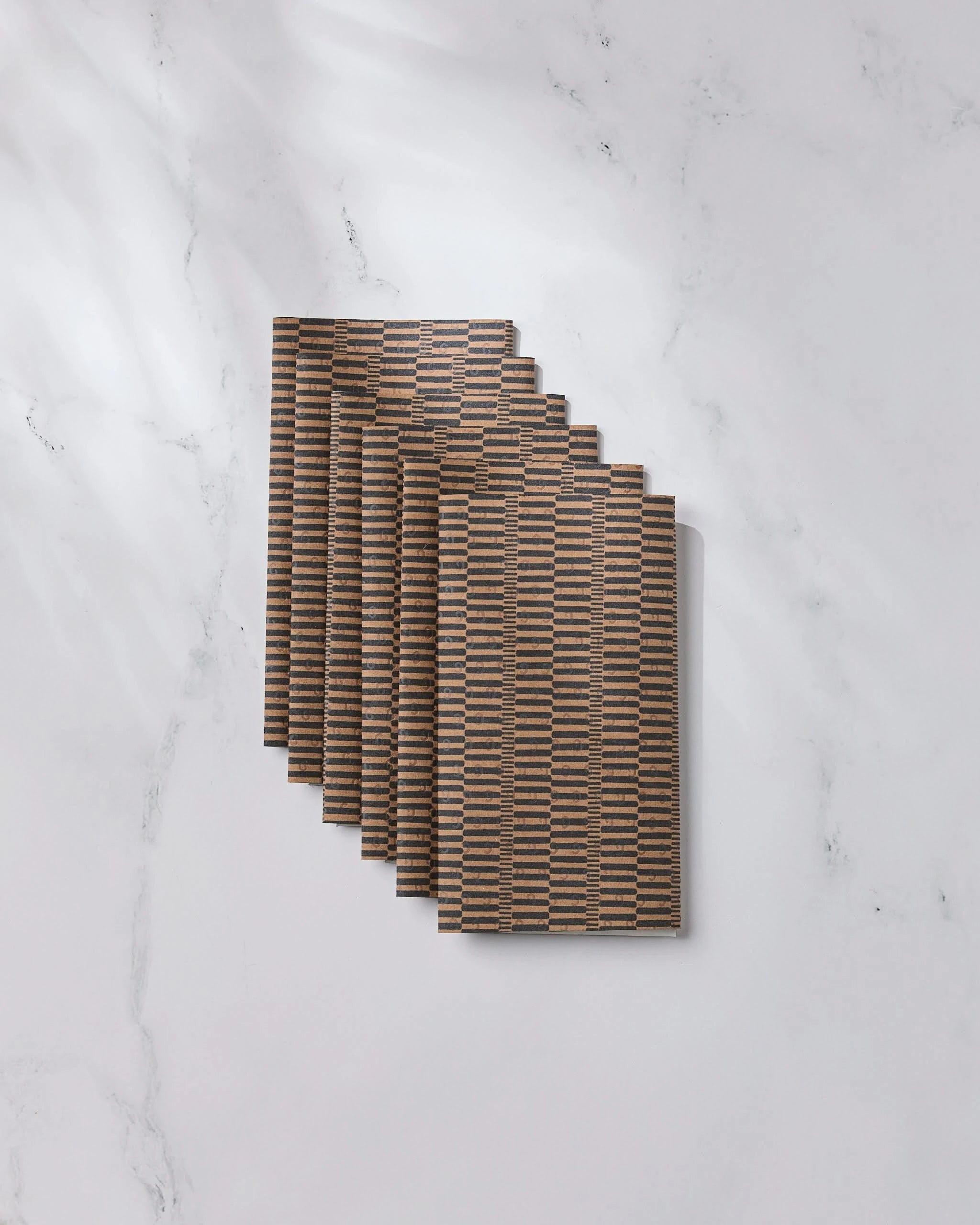 Geometry’s Ridge reusable paper towels, featuring brown and black stripes, are fanned out on a white marble surface with gentle lighting and shadows.