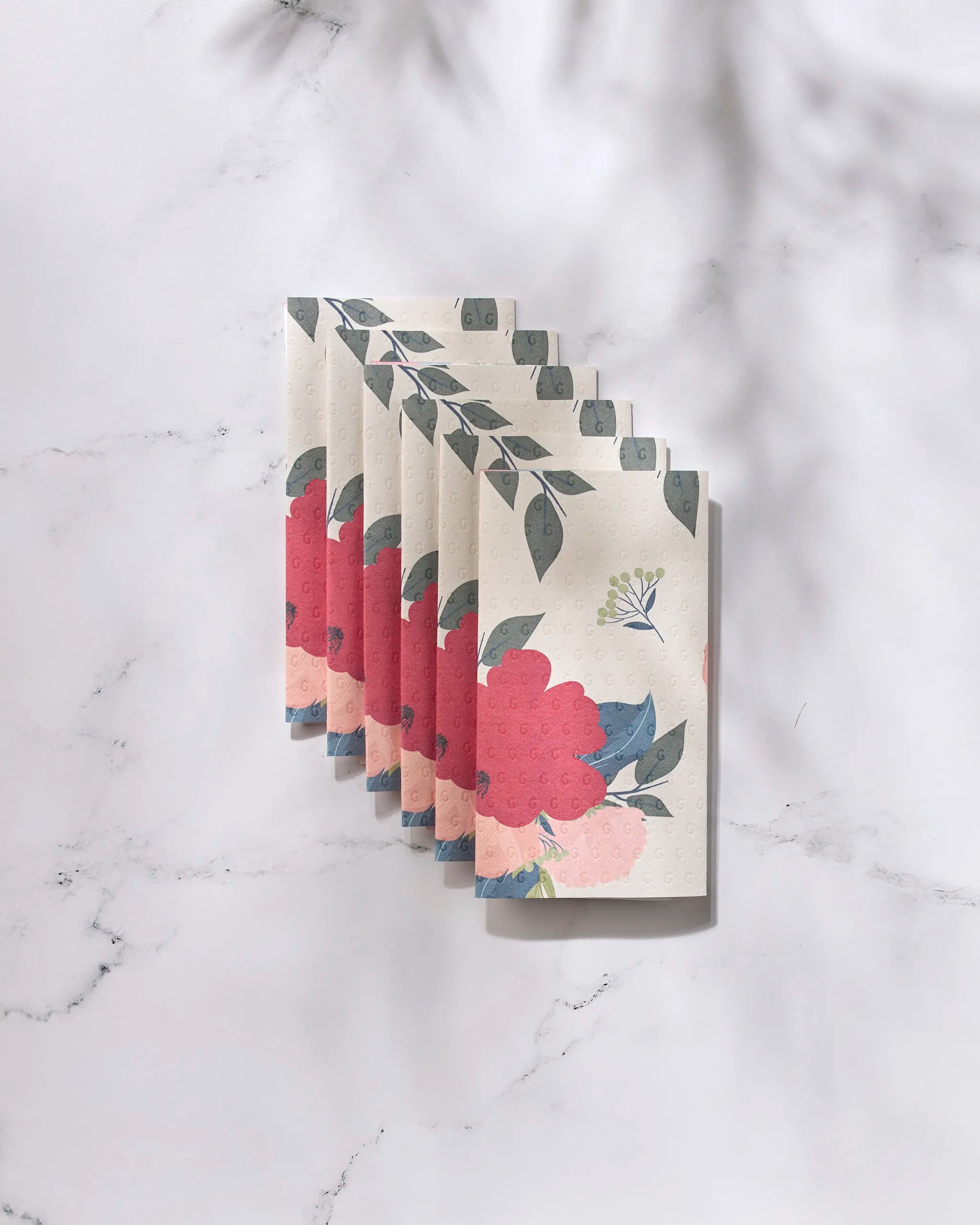 Five "Showered With Love" napkins by GEOMETRY, featuring pink flowers and green leaves, are fanned out on a white marble surface with soft shadows, blending eco-friendly elegance with floral charm.