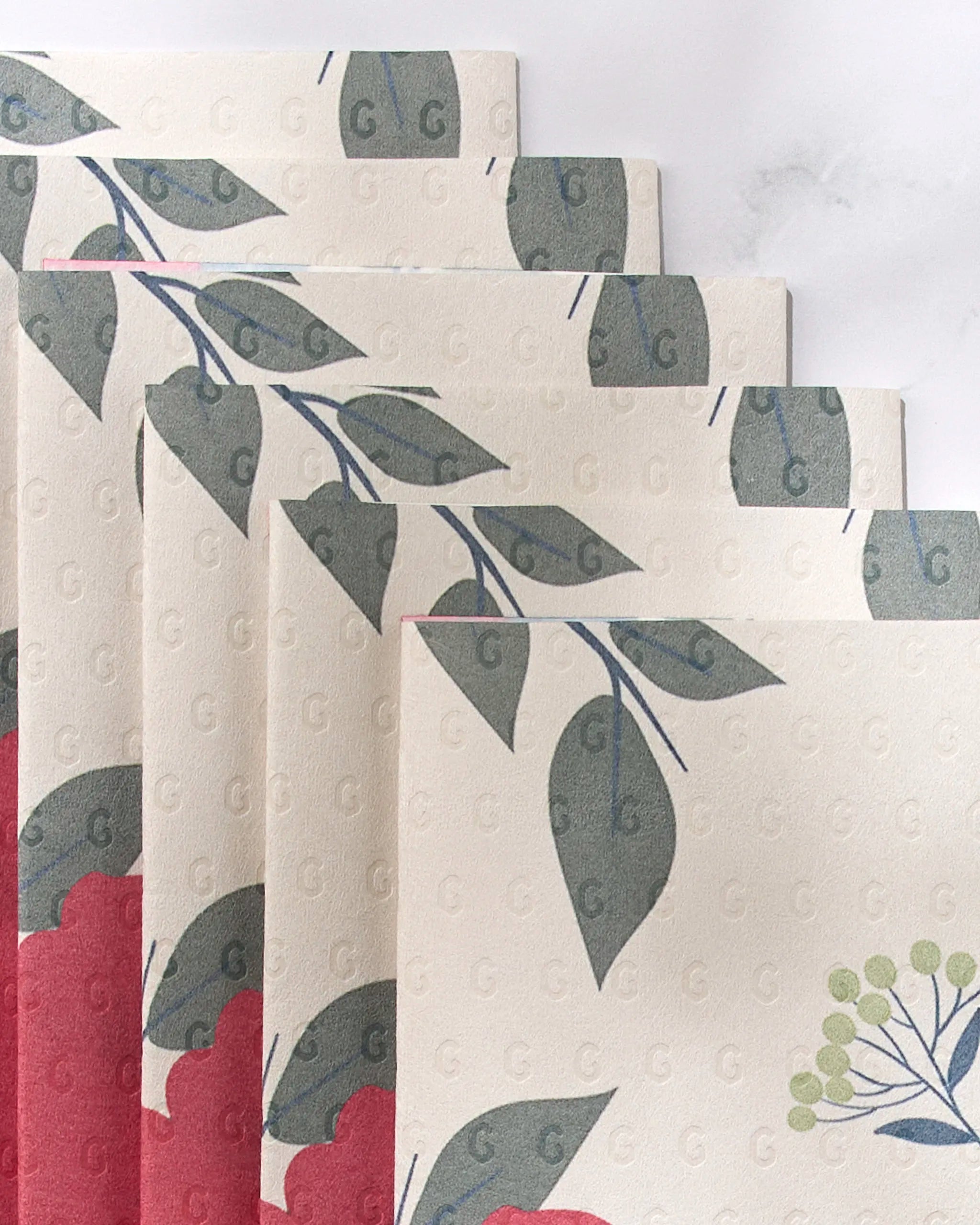 Five reusable paper towels from GEOMETRY’s "Showered With Love" collection are fanned out, featuring leafy green branches, red floral shapes, and light green clusters on a cream background embossed with subtle “G” letters.