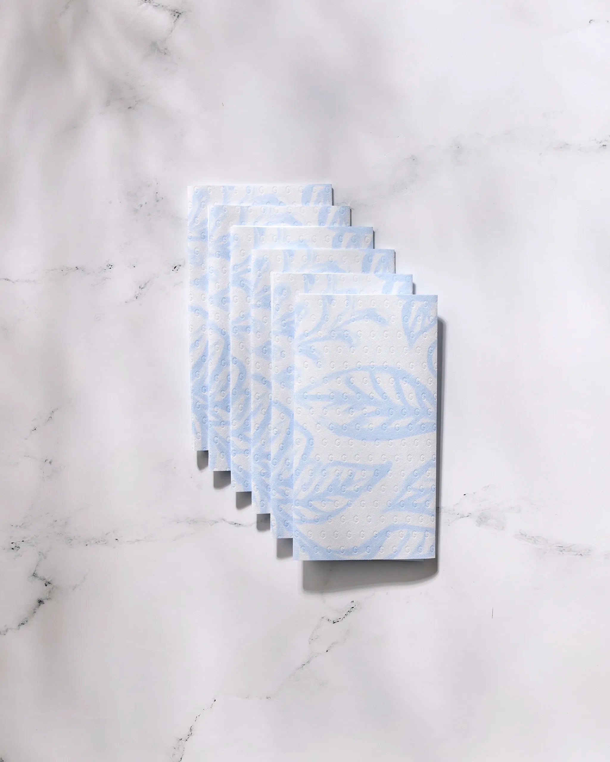 Six eco-friendly "Something Blue" paper towels by GEOMETRY, featuring a subtle blue leaf pattern, are neatly stacked on a white marble surface.