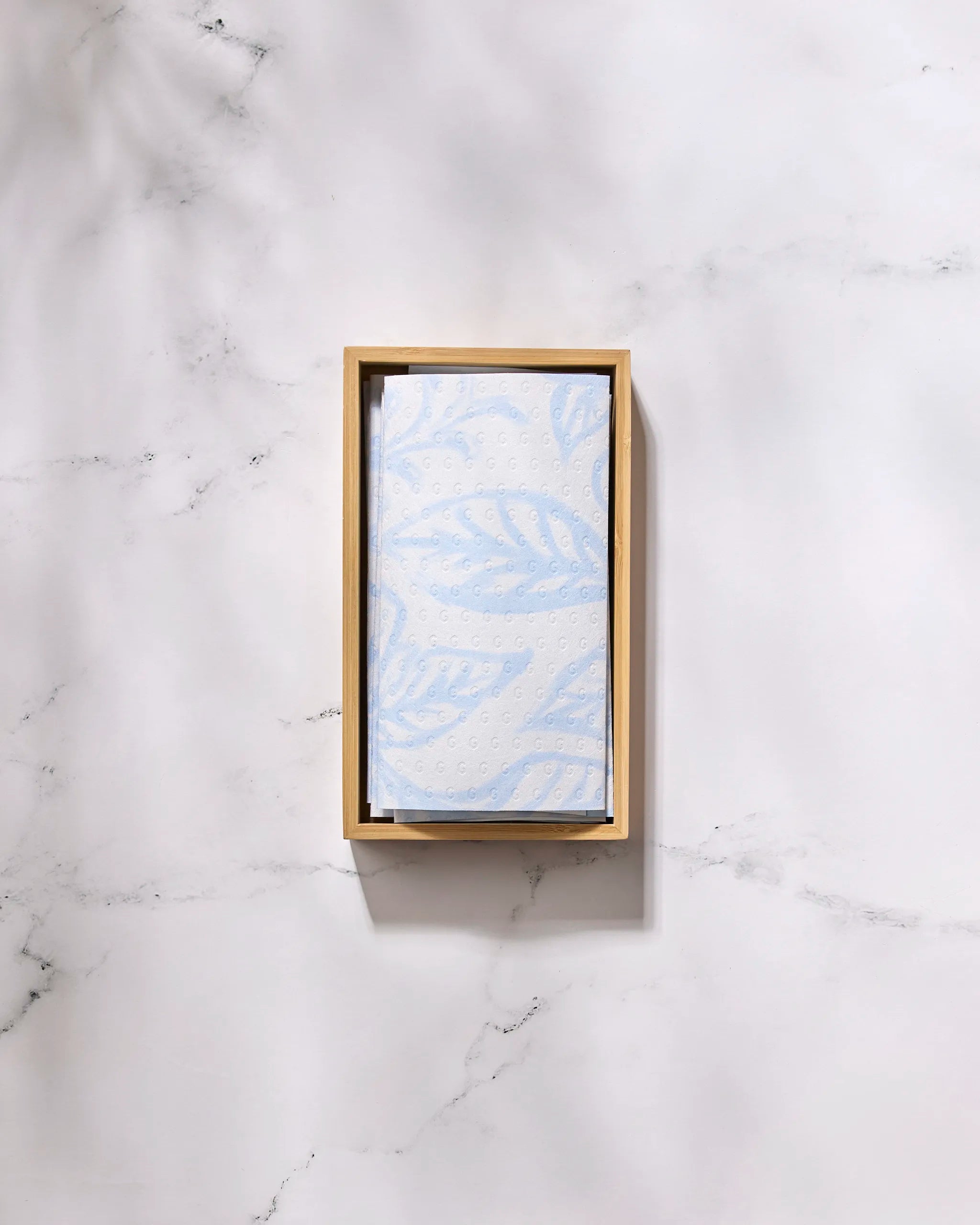 The GEOMETRY Something Blue eco-friendly, light blue paper towel with a leaf pattern is folded in a rectangular wooden tray on a white marble surface.