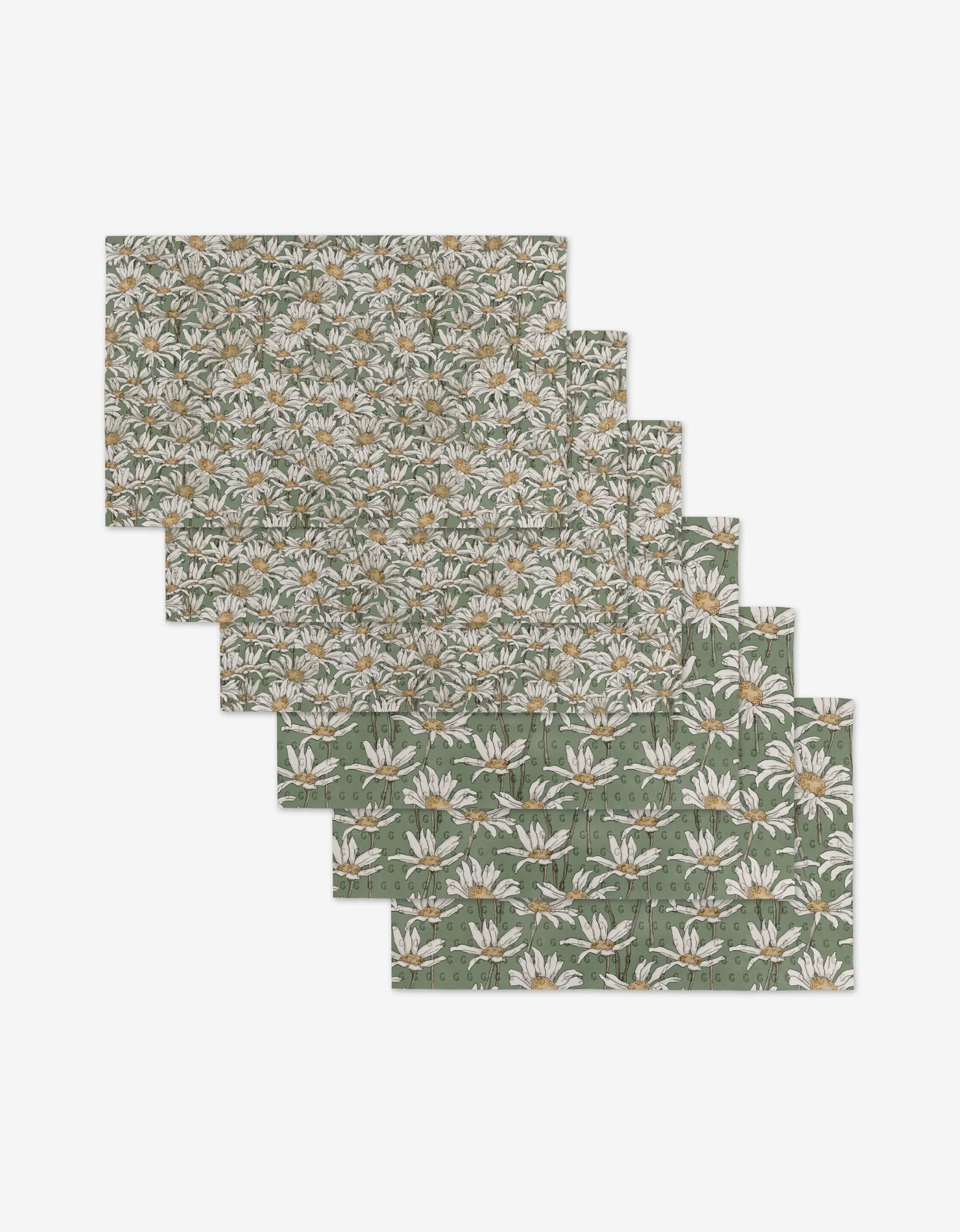 Six eco-friendly Aubrey Fairchild Springtime placemats, featuring a green base with white daisies and yellow centers, are fanned out to display their floral design. Theyre made from post-consumer recycled materials.