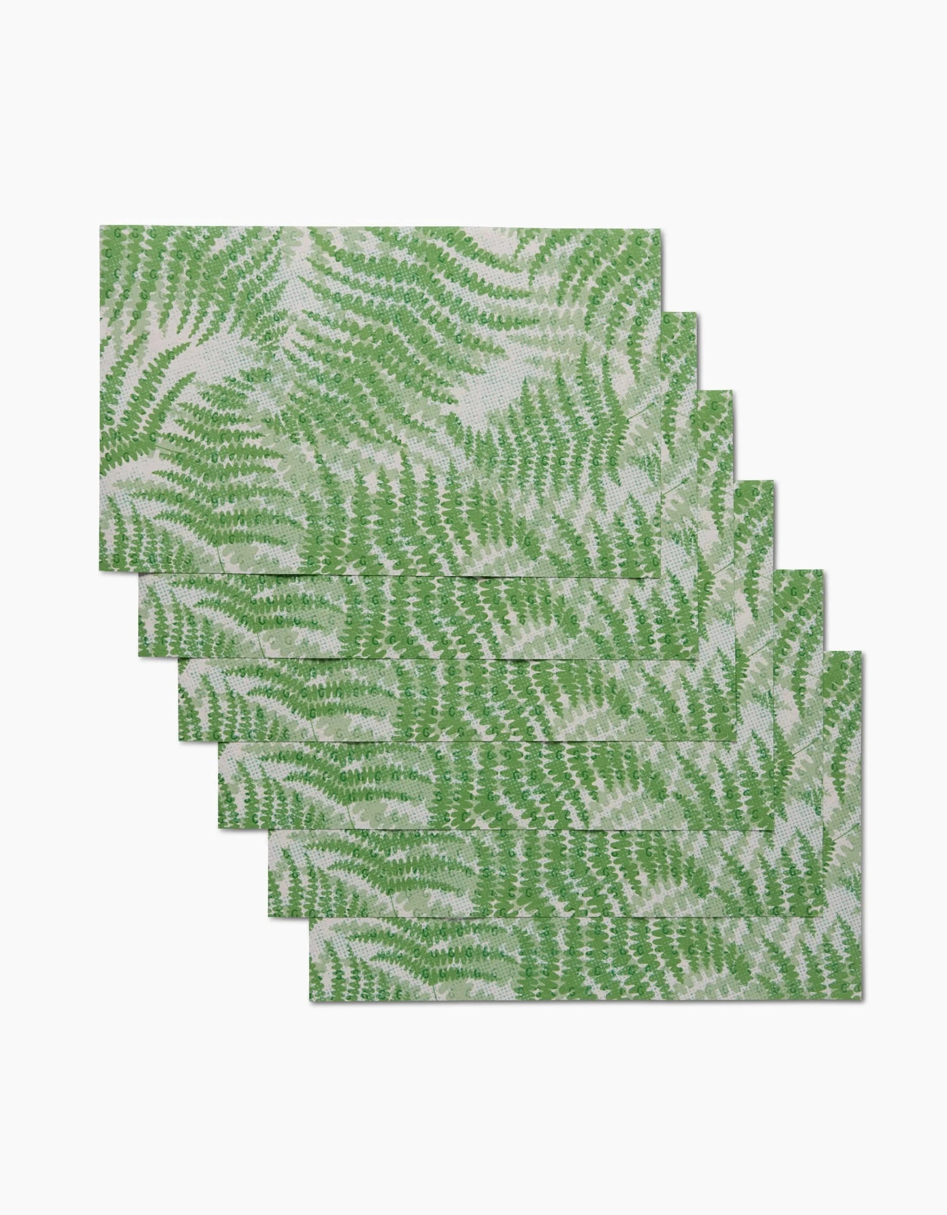 Six Swaying Leaf placemats by GEOMETRY, featuring a green fern pattern on white and made from eco-friendly materials, are shown stacked in a staggered arrangement on a white surface.