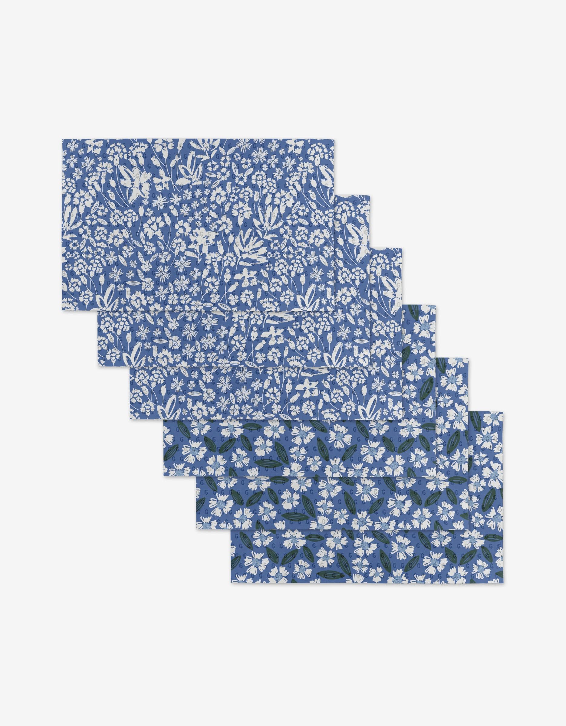 Six Sweetie Floral rectangular placemats by Hufton Studio are fanned out, showing blue backgrounds with white floral patterns. Made from recycled materials, the bottom placemats feature a darker blue and green accents.