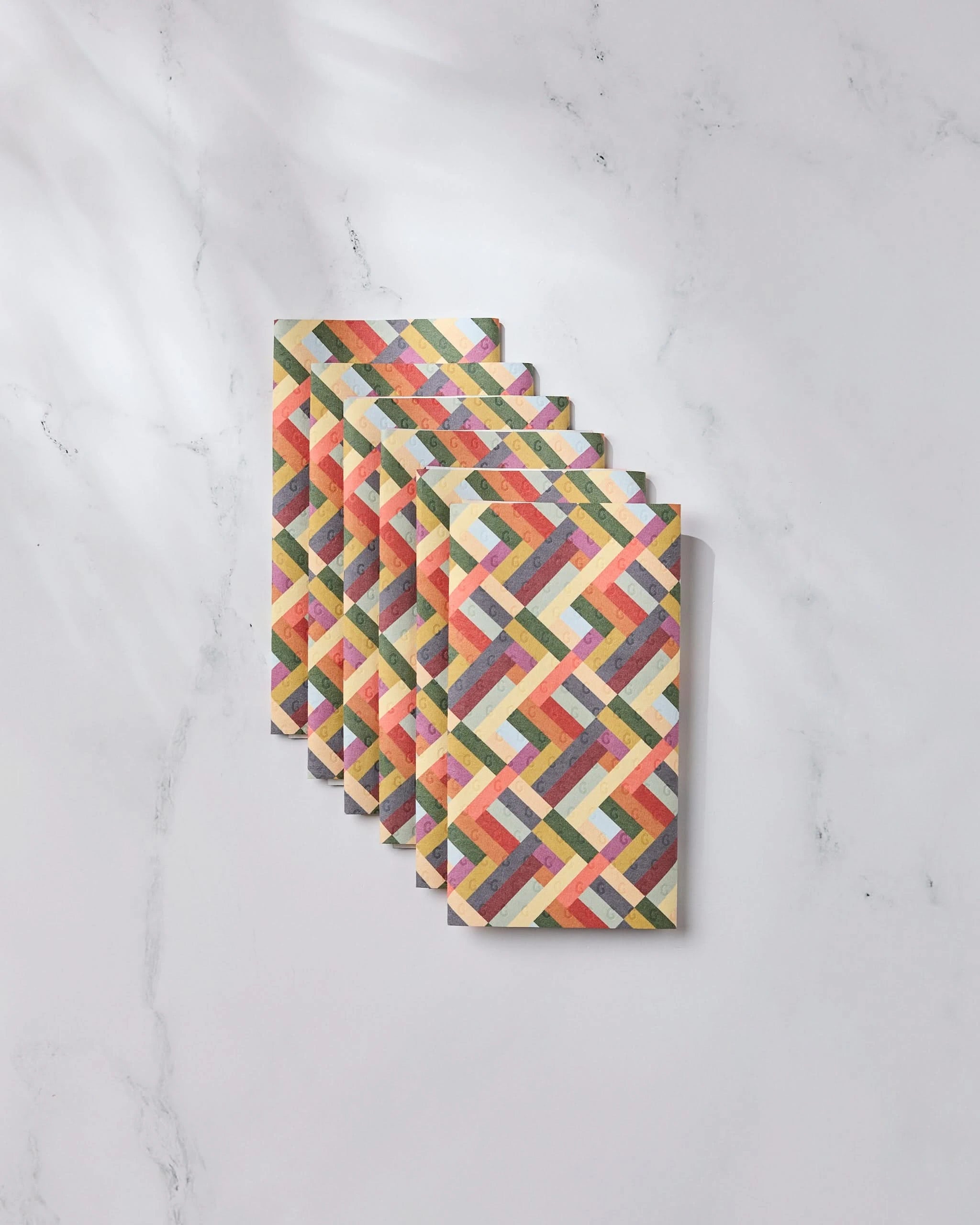 A stack of Vivid Cascade notebooks by Geometry, featuring colorful, geometric zigzag covers, is arranged on white marble—evoking the eco-friendly vibrance of reusable paper towels.