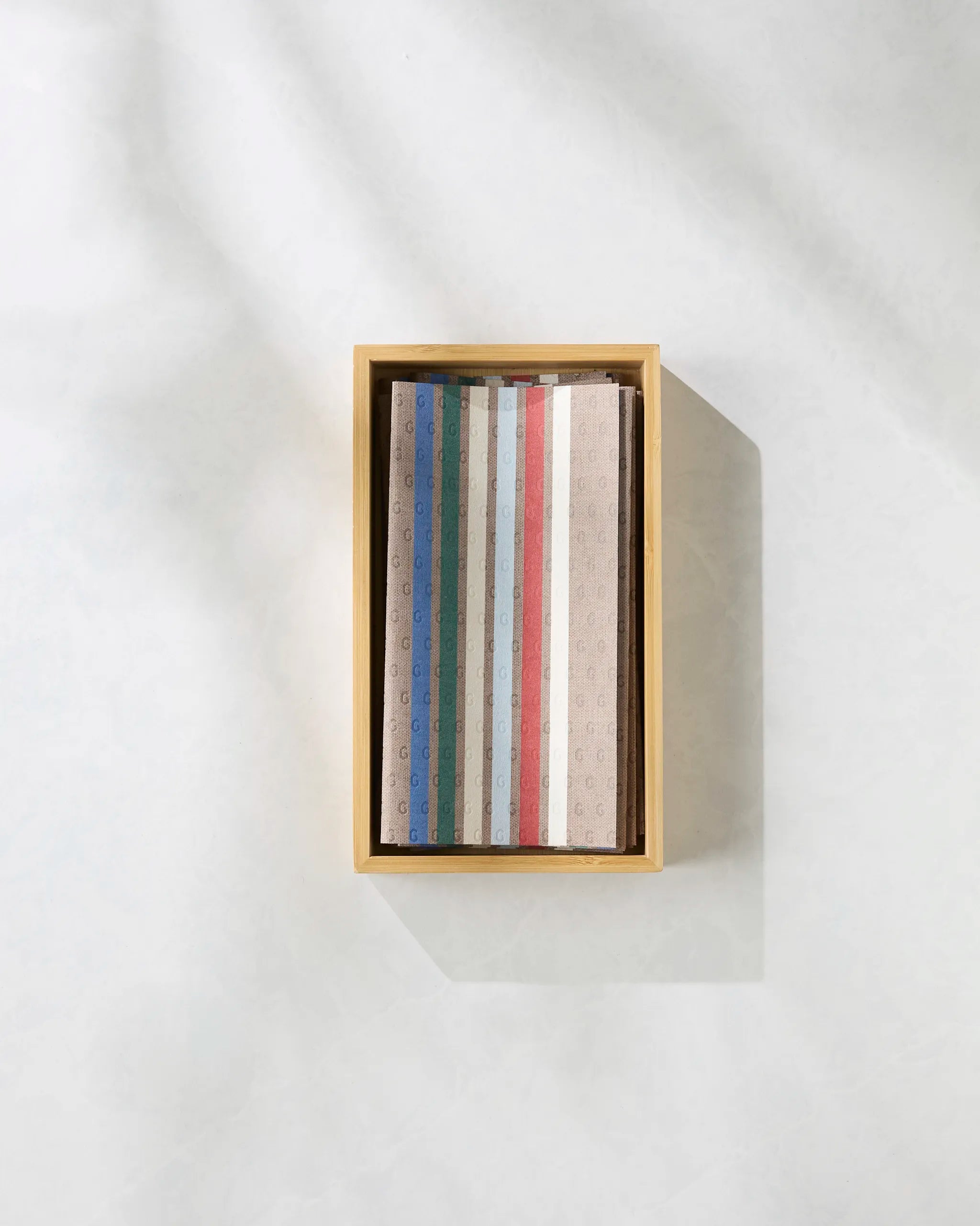 The Winterhaus by Geometry is a wooden box with neatly stacked, colorful patterned sheets—blue, green, red, and beige—seen from above on a light surface, offering an eco-friendly alternative to Not Paper Towels.