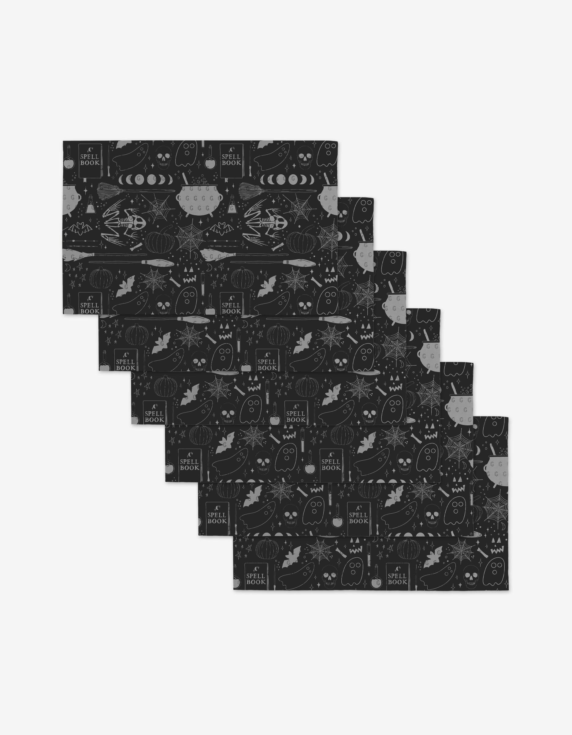 Six black rectangular “Witches Brew” placemats by Rosa Kichline, featuring white autumn motifs and “Hello Fall,” are fanned out. Made from eco-friendly materials for a sustainable alternative to paper towels.