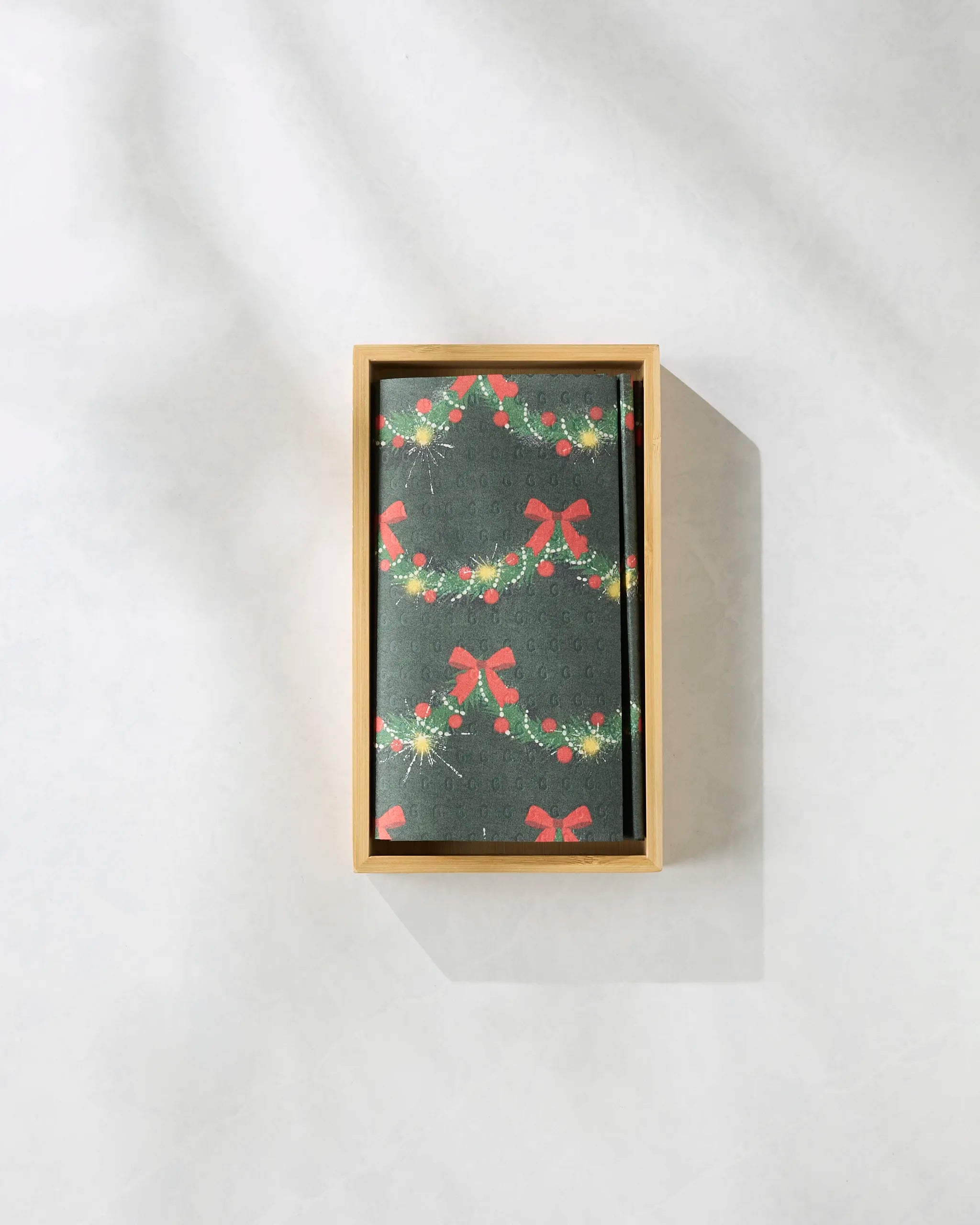 The Geometry "Yuletide Garland" notebook, featuring festive red bows and green garlands on the cover, rests in a wooden box against a light gray background—an ideal gift with eco-friendly kitchen towels.