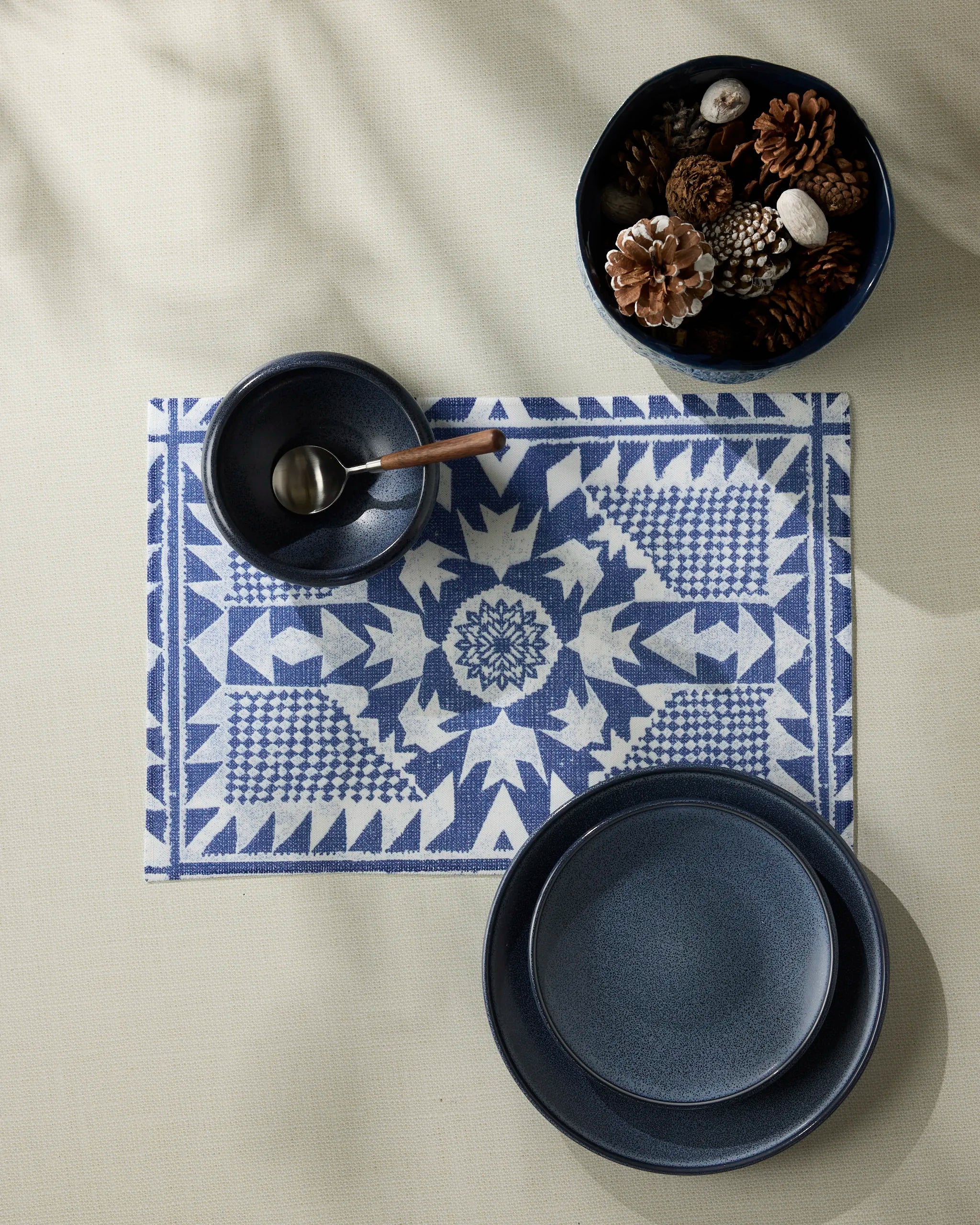 The Geometry Bluebird placemat features blue and white patterns, paired here with a blue bowl, plate, small bowl with silver spoon, and a bowl of pinecones on a light tablecloth. Stylish and machine washable for easy care.