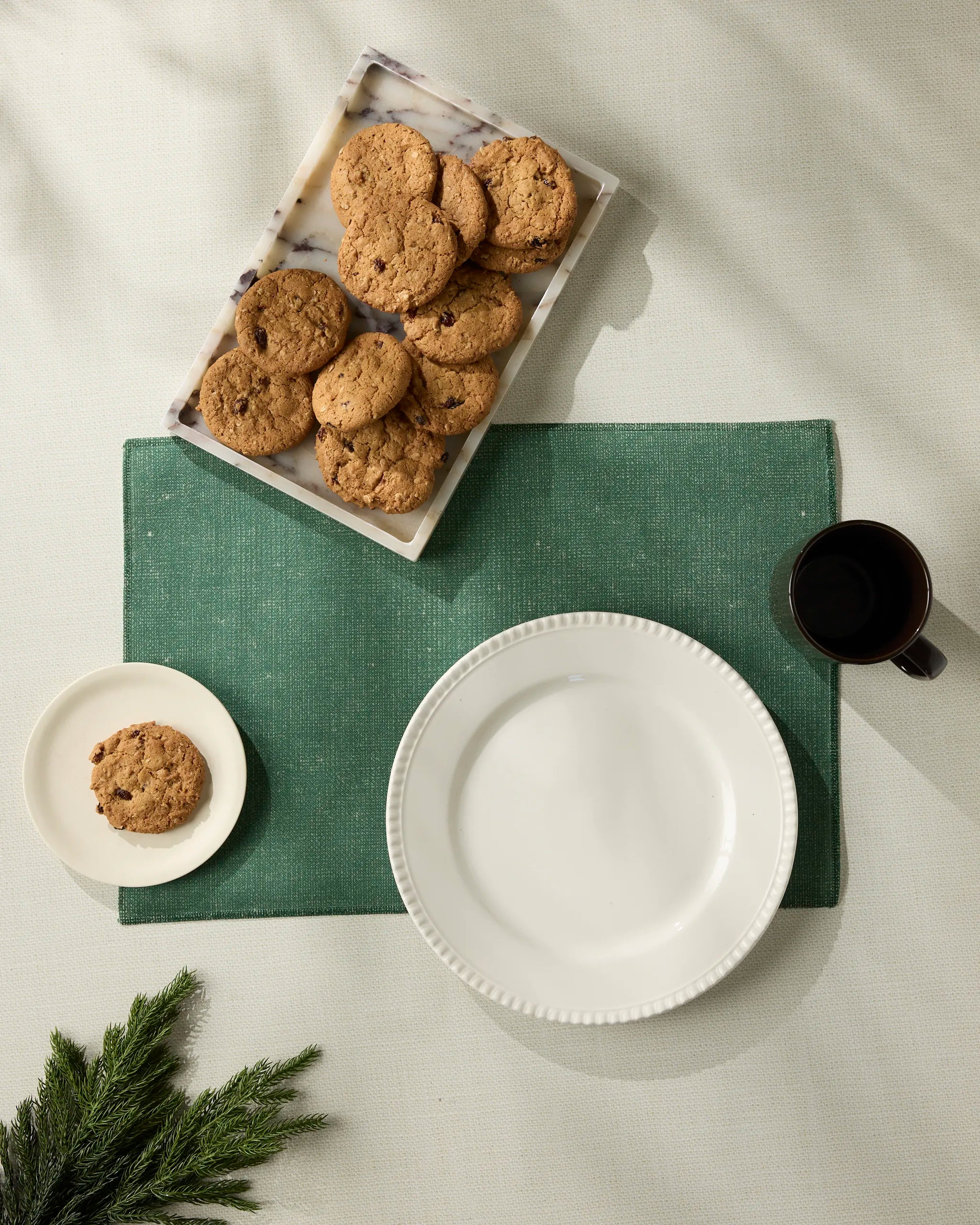 Overhead view of a table featuring the Geometry Cedar green machine-washable placemat, a large white plate, a small white plate with a cookie, a tray of cookies, a black mug, and green foliage in the corner.