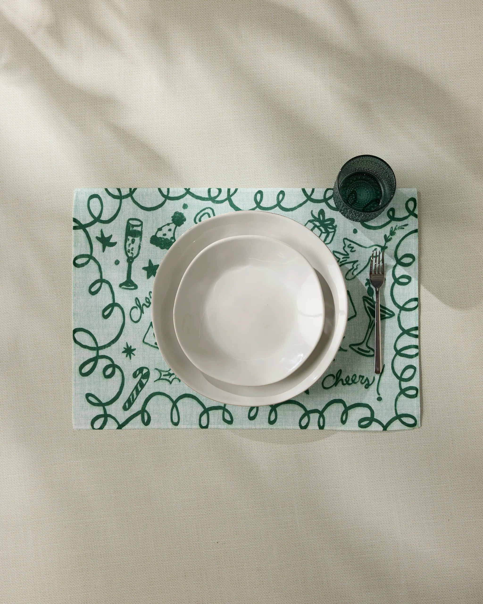 A white ceramic bowl and plate sit atop a green and white “Doodles for Holiday” placemat by Geometry, featuring celebratory doodles and “Cheery,” beside a fork and dark green glass. The placemat is machine washable.