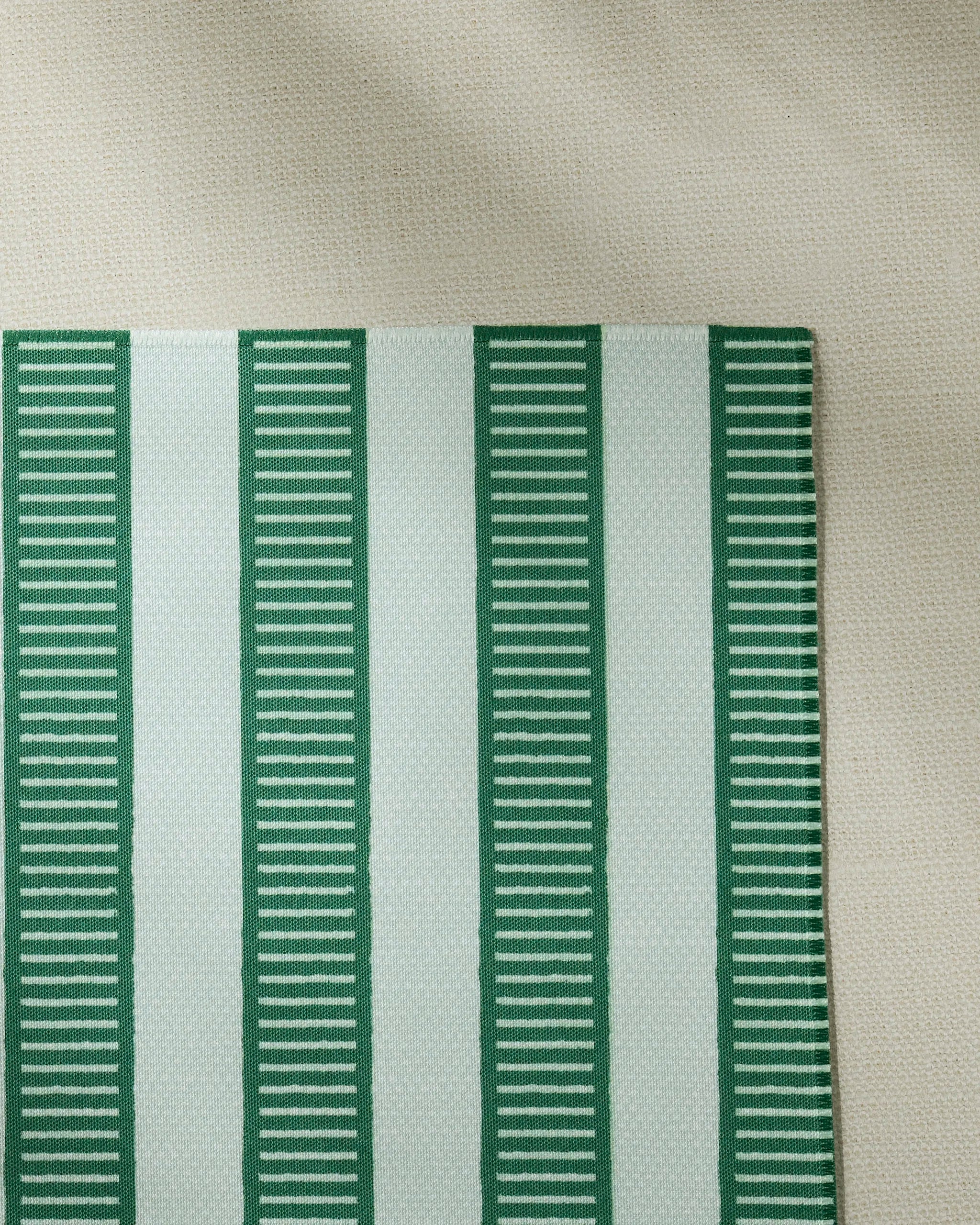 The Geometry Firstripe recycled rectangular placemat, featuring alternating vertical green and white stripes with horizontal green dashes, is shown on a textured beige surface.
