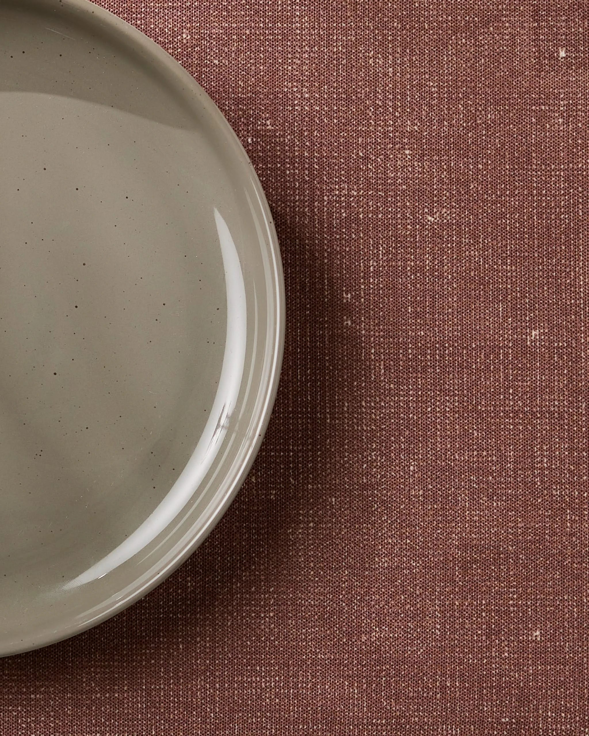 A close-up of Geometry’s eco-friendly Russet placemats showcases their style on a textured, reddish-brown background beside a light gray ceramic plate, which is partially visible and empty on the left.