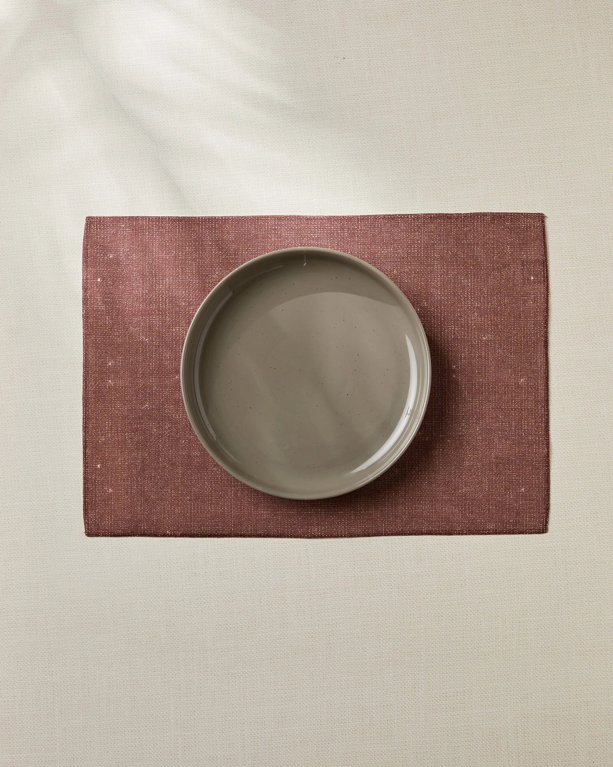 The Geometry Russet eco-friendly placemat in maroon, with a beige ceramic plate on top, rests on a light tablecloth, captured from above for a simple, minimalistic look.