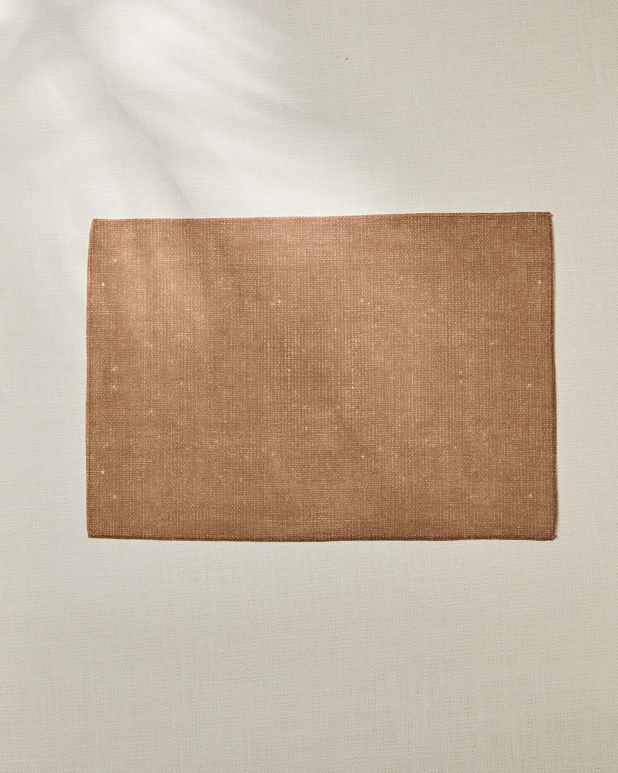 The Geometry Wheat rectangular brown woven mat is displayed on a light beige textured surface, with soft natural light creating subtle shadows in the top left corner.