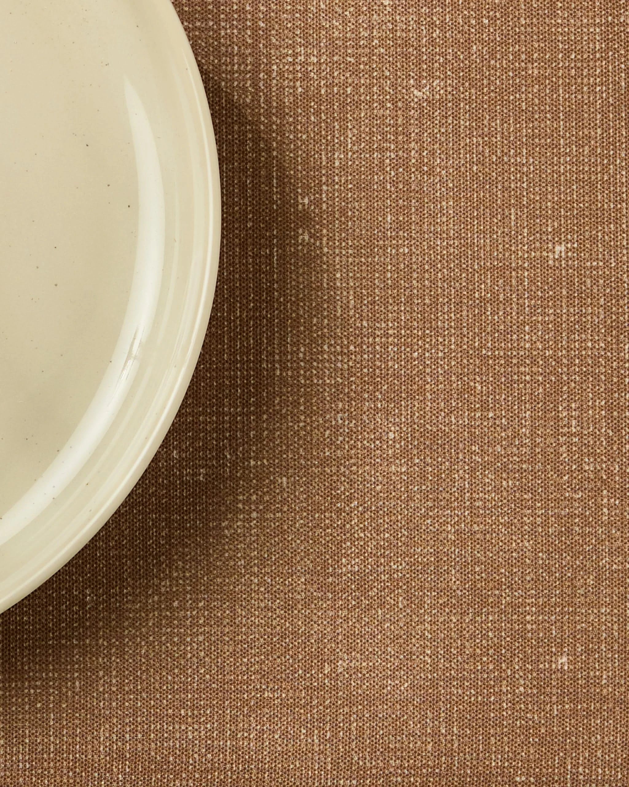 A close-up of the edge of a cream-colored ceramic plate on Geometrys Wheat placemat, with the plate in the upper left corner atop textured brown fabric.