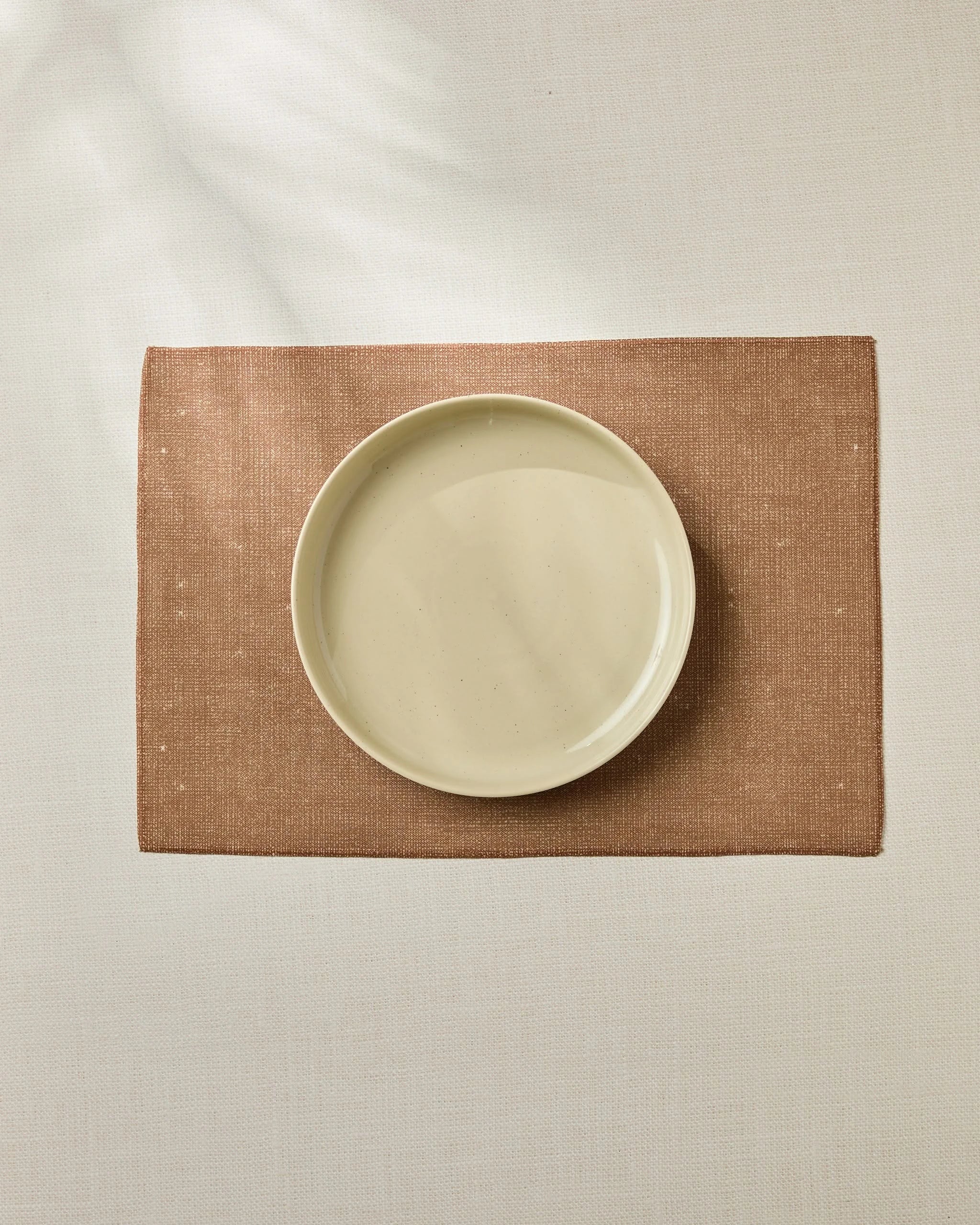 A beige Wheat ceramic plate by Geometry is centered on a brown, durable placemat atop a light-colored, textured surface.