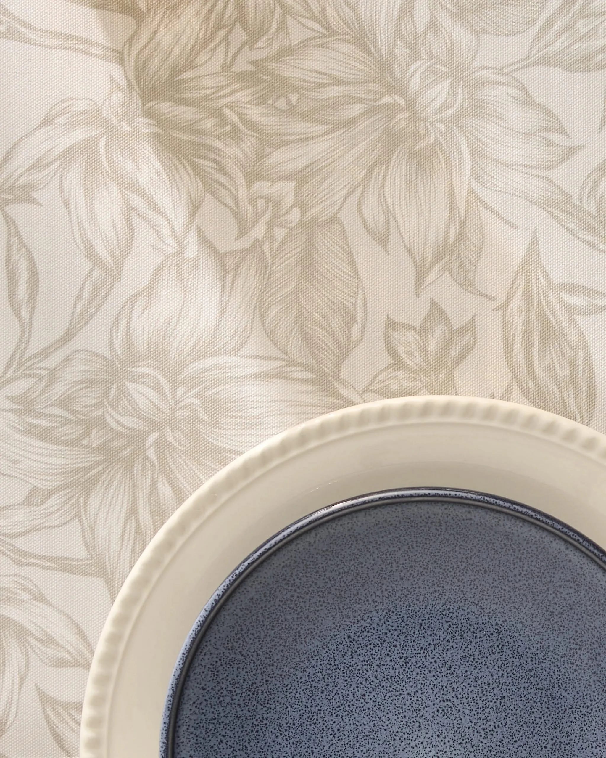 A close-up of the Alba blue ceramic bowl by Geometry, nestled in a cream-colored plate and set atop beige floral-patterned recycled placemats.