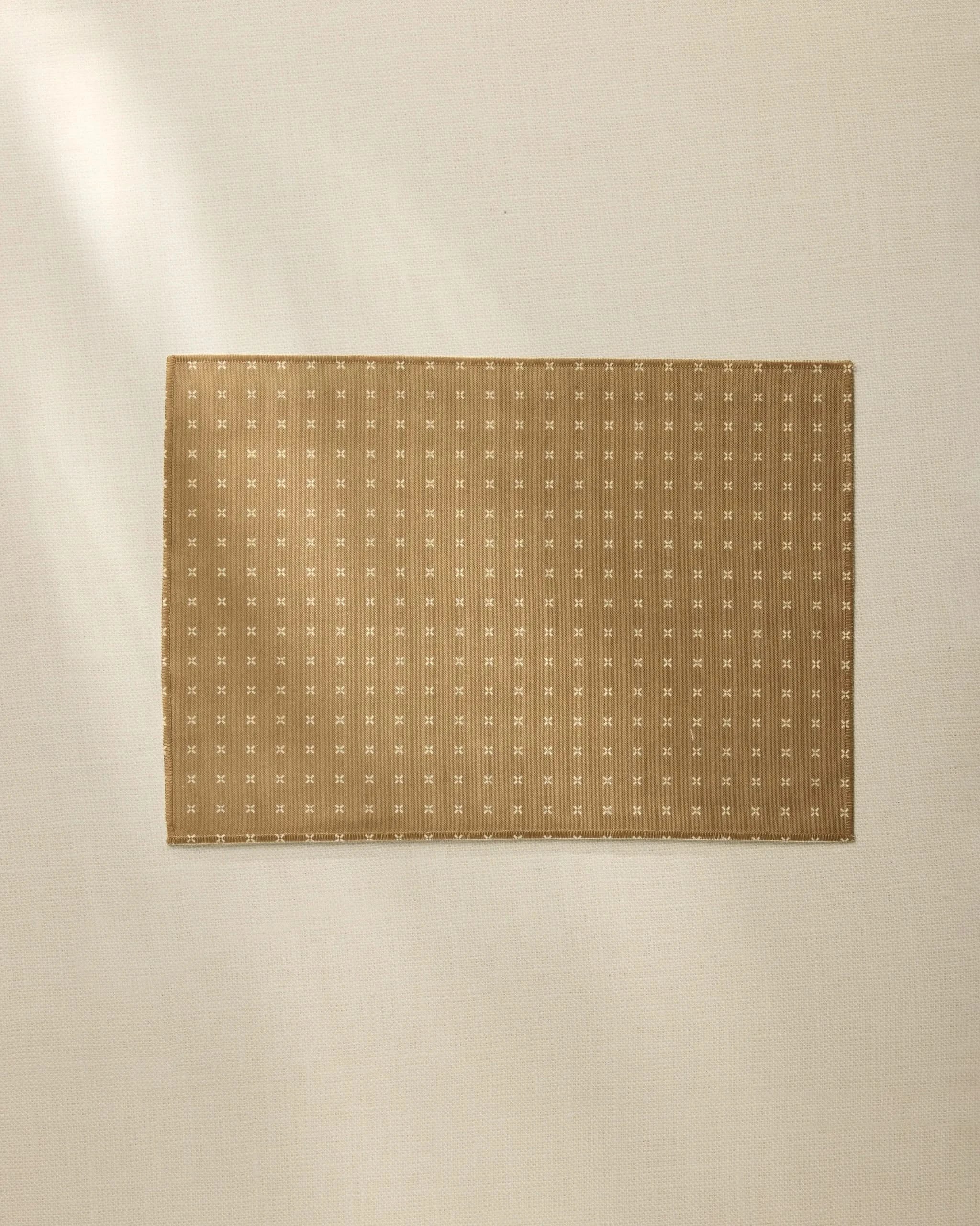 The Amber Stitch placemat by Geometry features a rectangular brown design with a small white cross pattern, lying flat on a light beige surface. Soft natural light highlights its style—machine washable and perfect for inviting table settings.