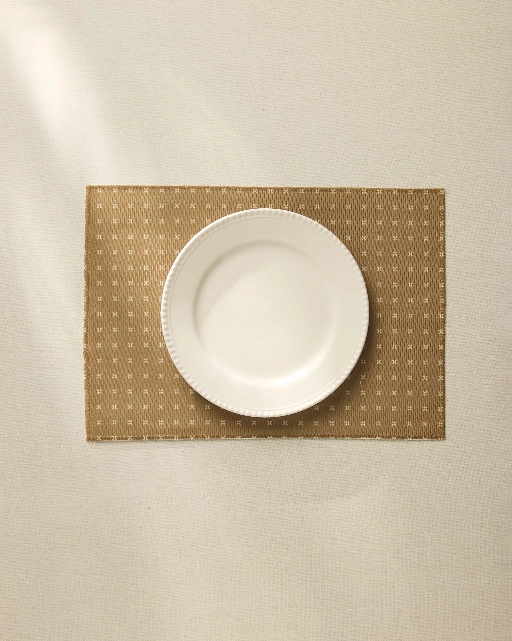 A white plate sits centered on a Geometry Amber Stitch Placemat, which rests on a light, textured tablecloth and is softly illuminated by natural light.