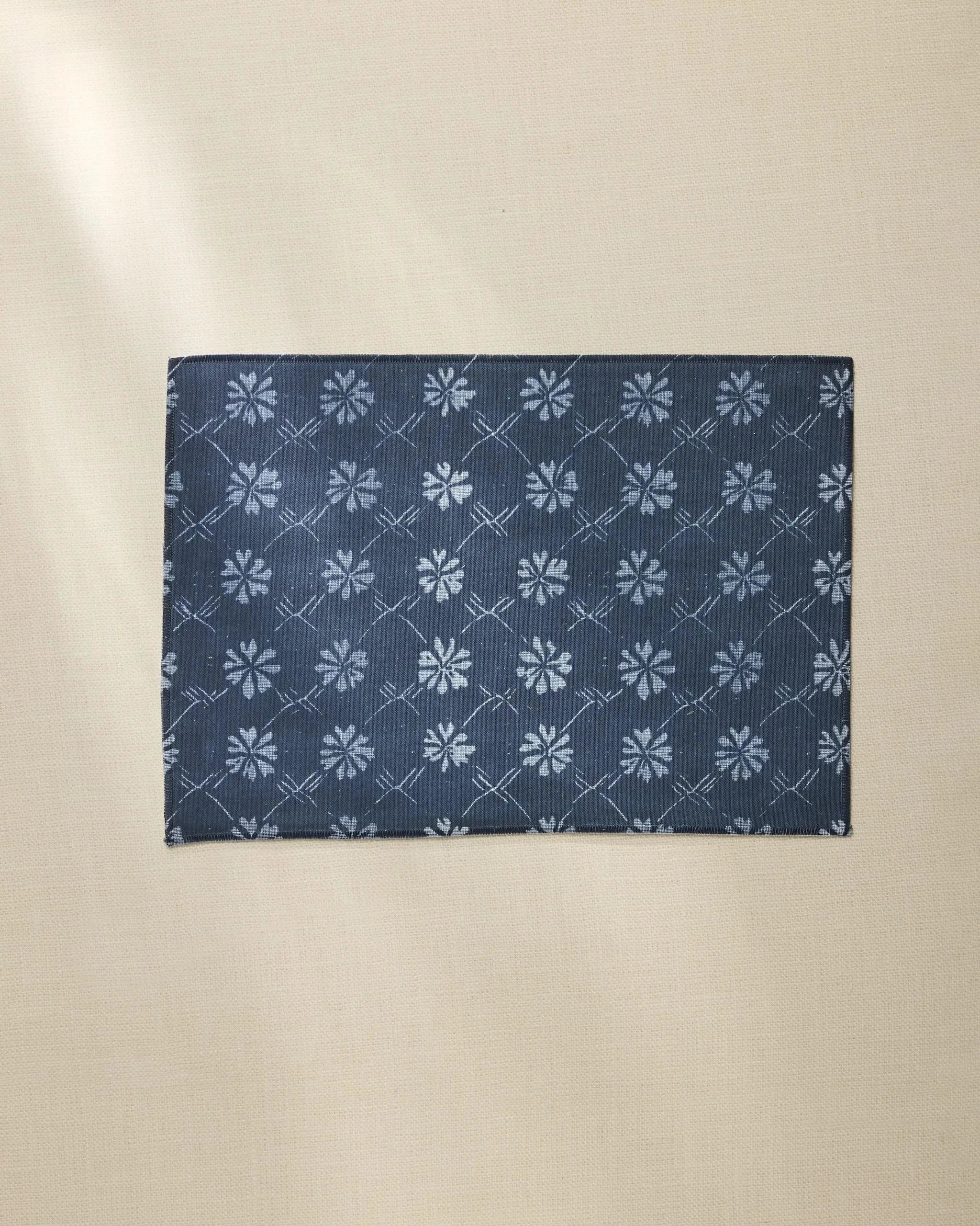The Geometry Bonnie Indigo rectangular placemat, featuring a dark blue color and subtle light blue floral pattern, rests on a beige textured surface.