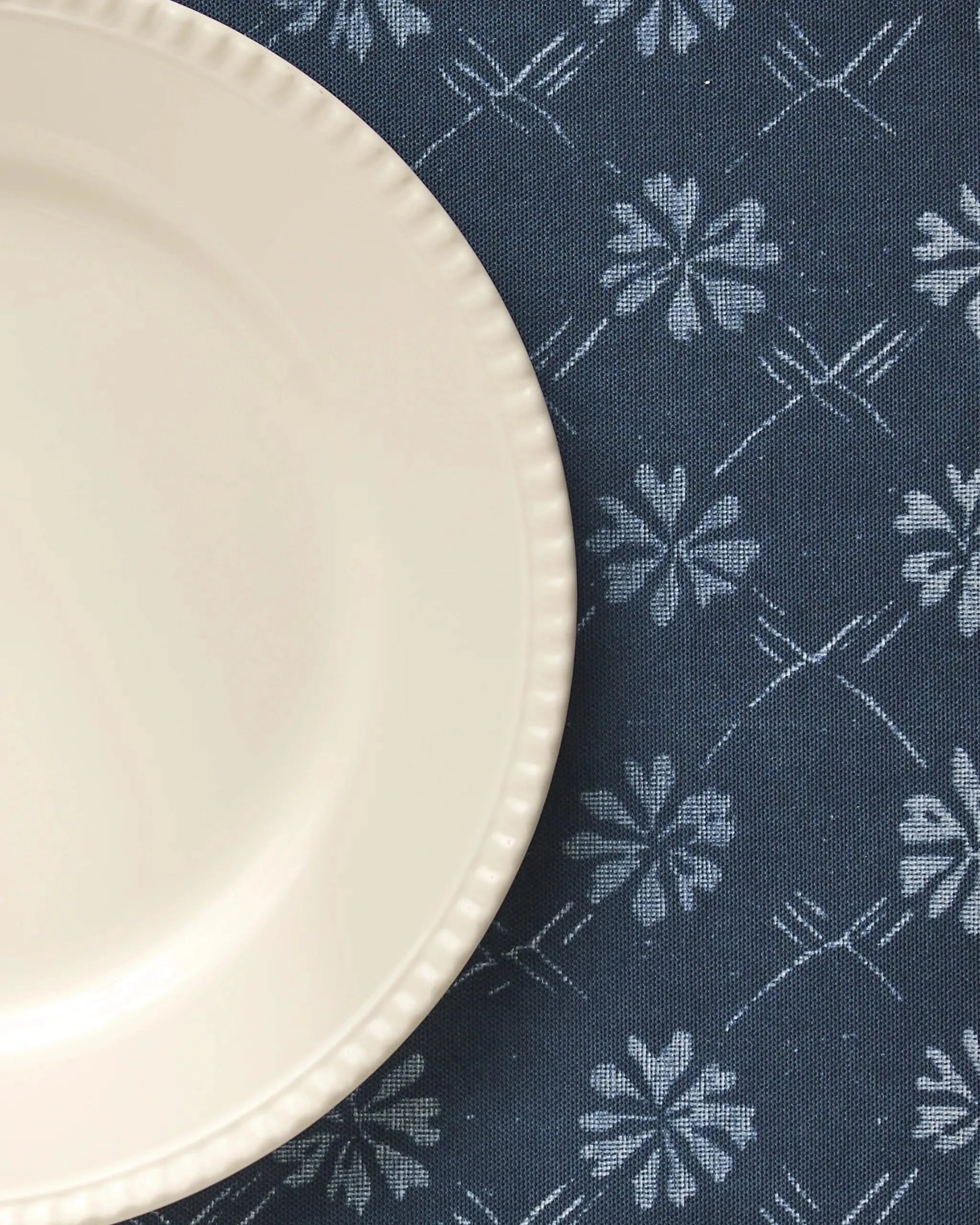 A cream-colored plate with a scalloped edge sits on the left side of a dark blue Geometry Bonnie Indigo placemat, featuring white floral and geometric patterns.