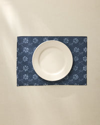 A white ceramic plate is centered on the Geometry Bonnie Indigo machine washable placemat, featuring a light floral pattern, atop a cream-colored tablecloth.