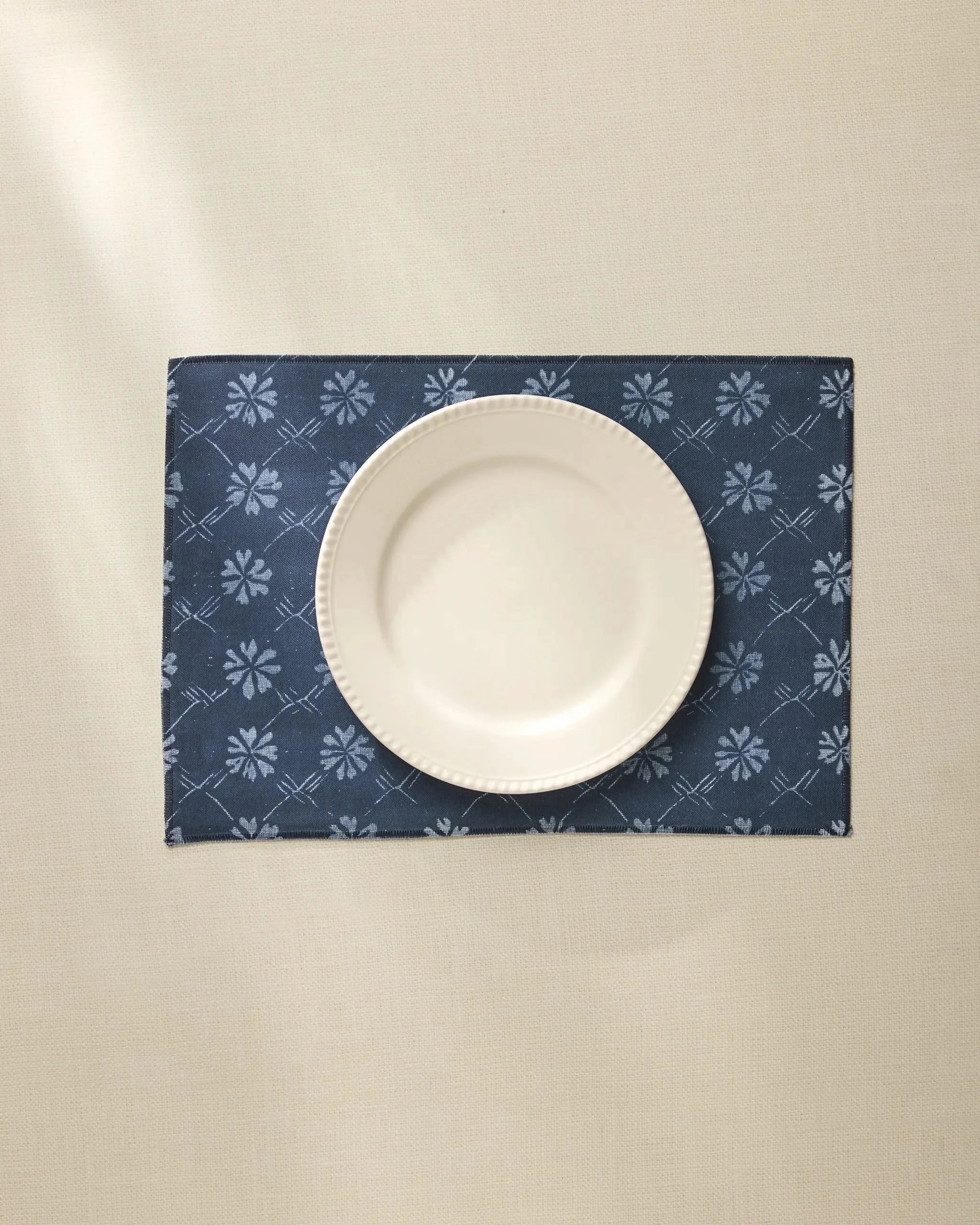 A white ceramic plate is centered on the Geometry Bonnie Indigo machine washable placemat, featuring a light floral pattern, atop a cream-colored tablecloth.