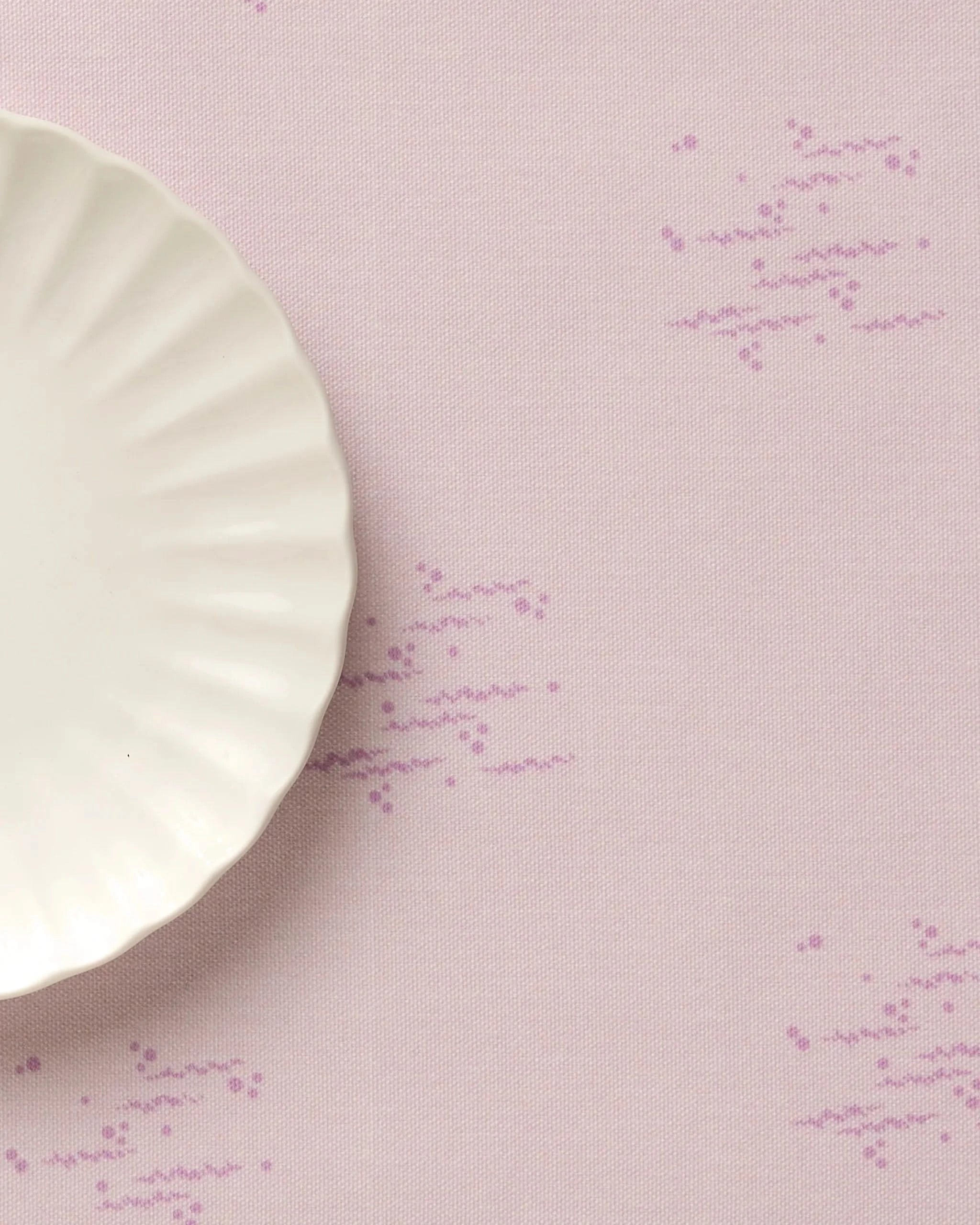 A white, scalloped-edge plate rests on a Bubble Ditzy Dusty Rose placemat by Geometry, featuring a dusty rose hue with scattered purple dots. Only part of the plate is visible in the upper left corner.