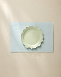 A light green, wavy-edged ceramic bowl sits centered on the Geometry Chambray Light Blue placemat, which rests on a beige tablecloth under soft, natural light.