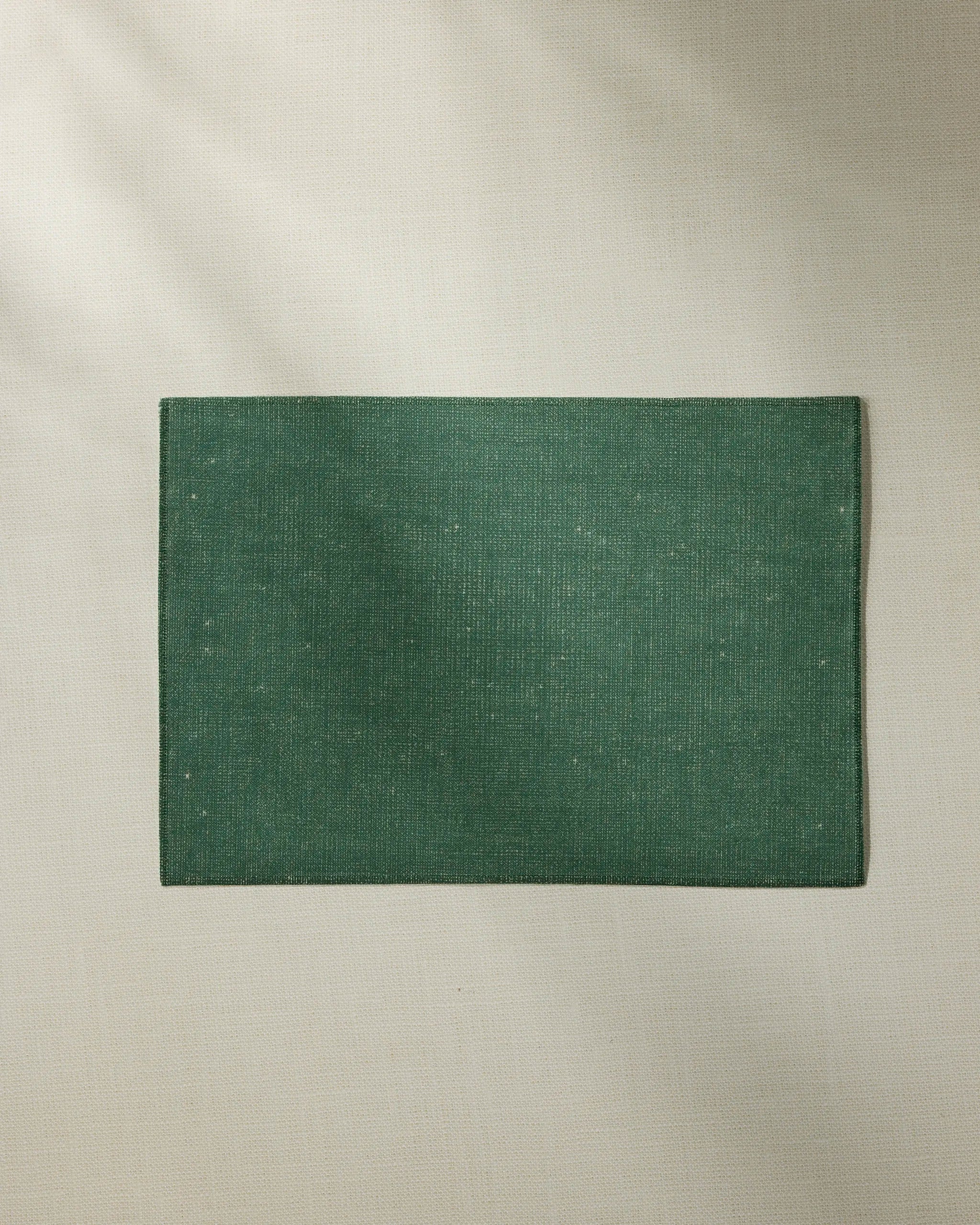 The Geometry Cedar placemat, in a rectangular green fabric, rests on a light beige textured surface with soft natural light and subtle shadows—an elegant, machine washable choice.