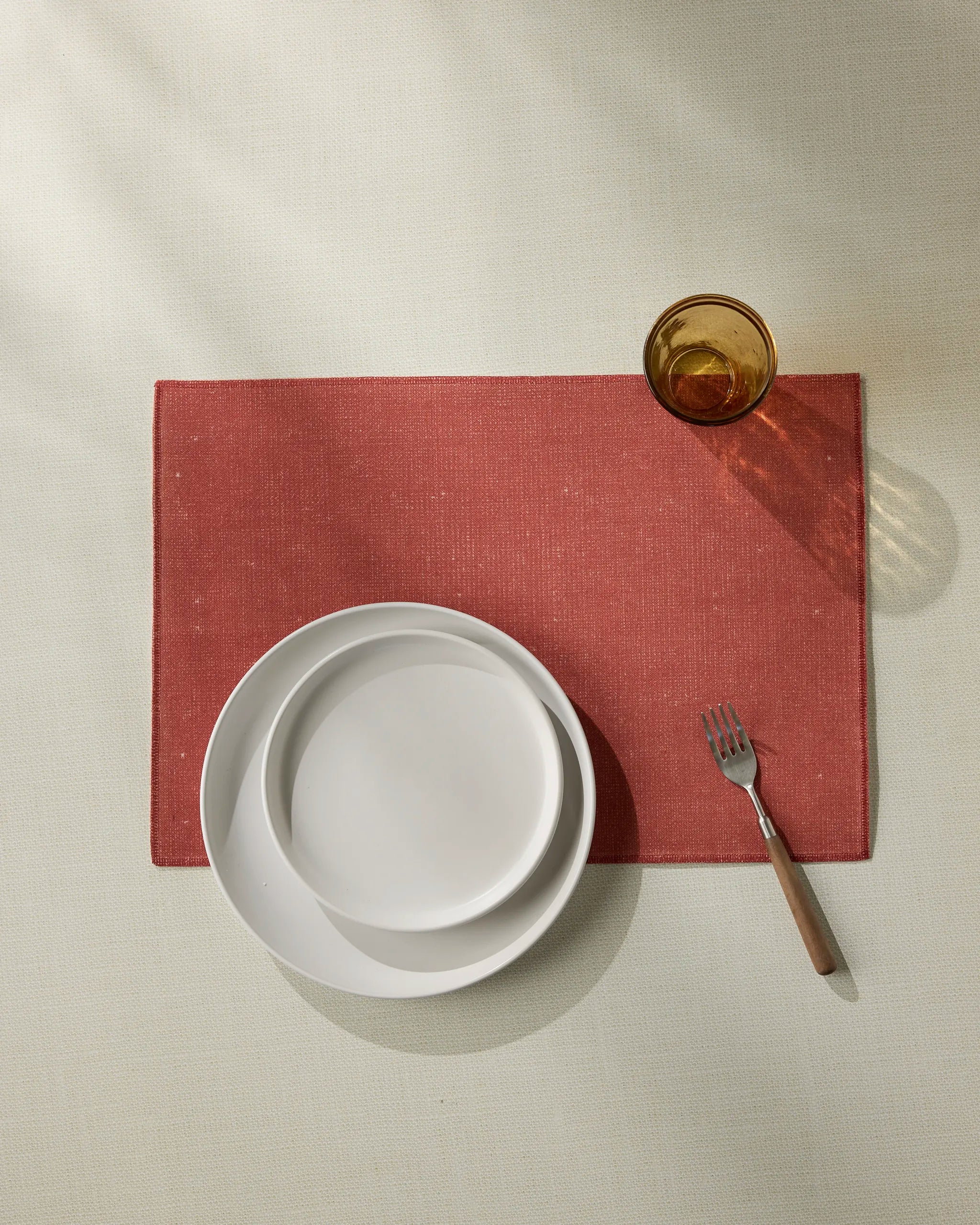 The Crimson placemat by Geometry sits on a light table, paired with two stacked white plates, a wooden-handled fork, and an amber glass casting a shadow. Machine washable for easy care.