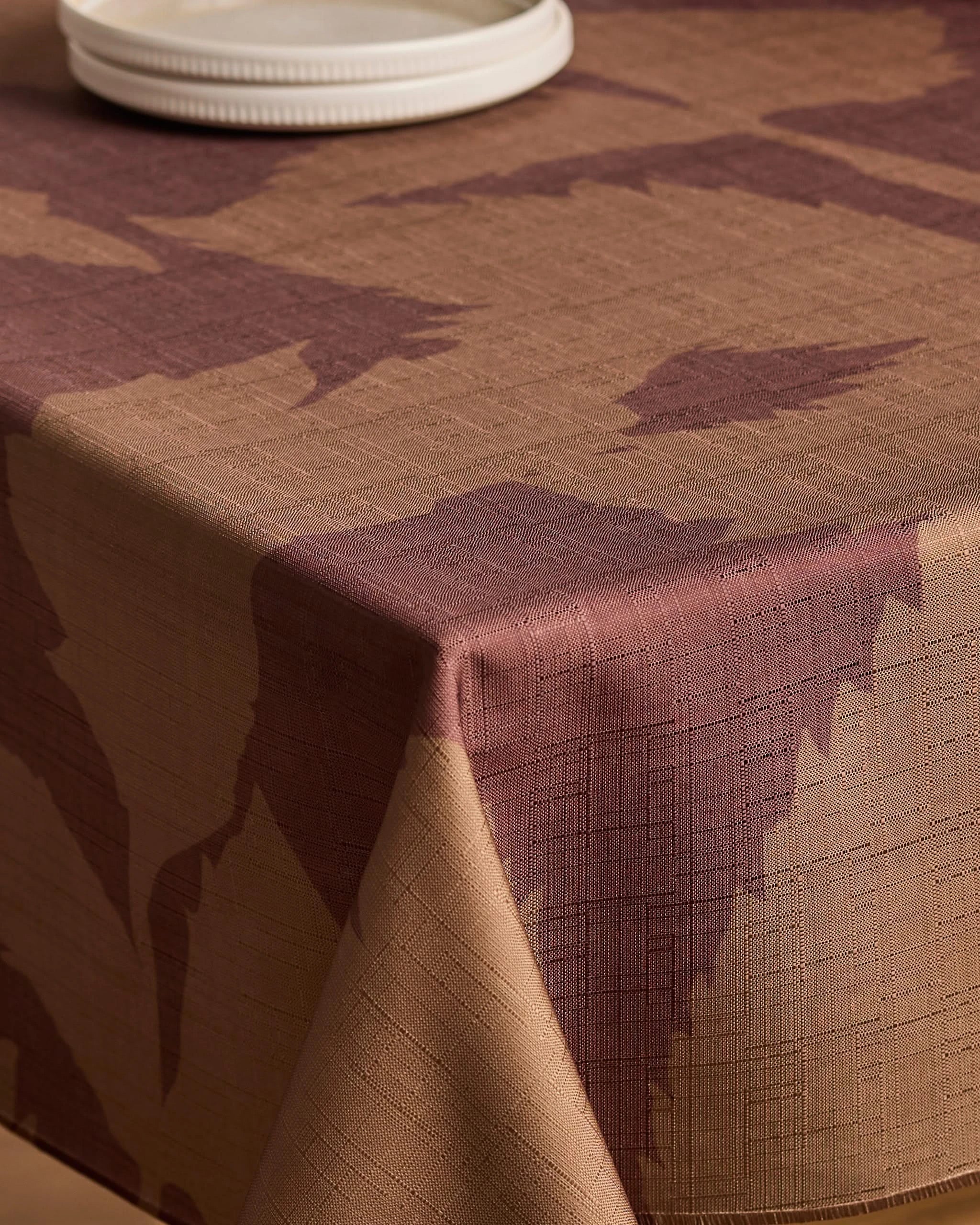 A close-up of the Geometry Rustle tablecloth, featuring brown and beige patterns with textured, stain-resistant fabric and crisp corners. White plates are stacked partially in view at the top.